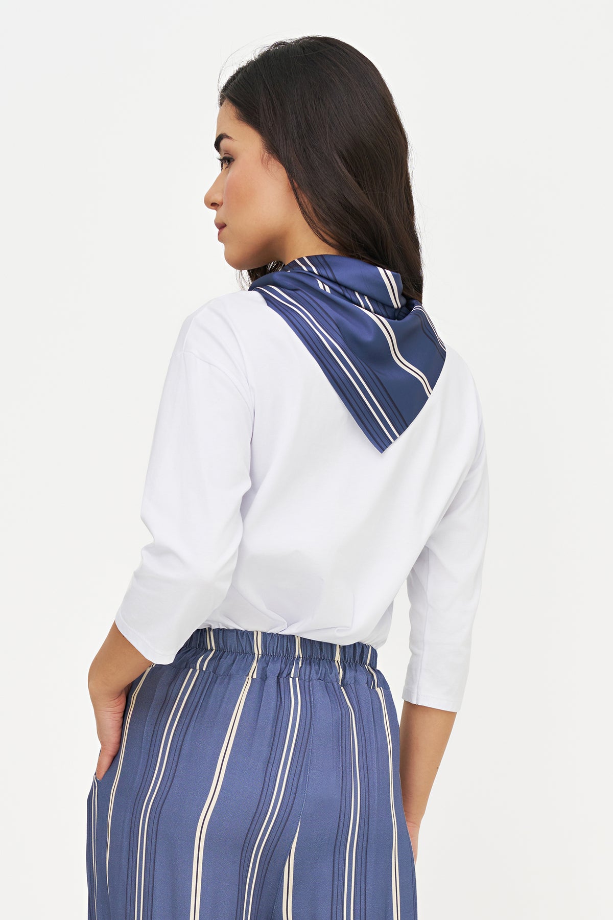 A woman shown from the back wearing a white blouse and blue striped trousers, with a matching blue striped scarf draped around her neck, against a plain light background.