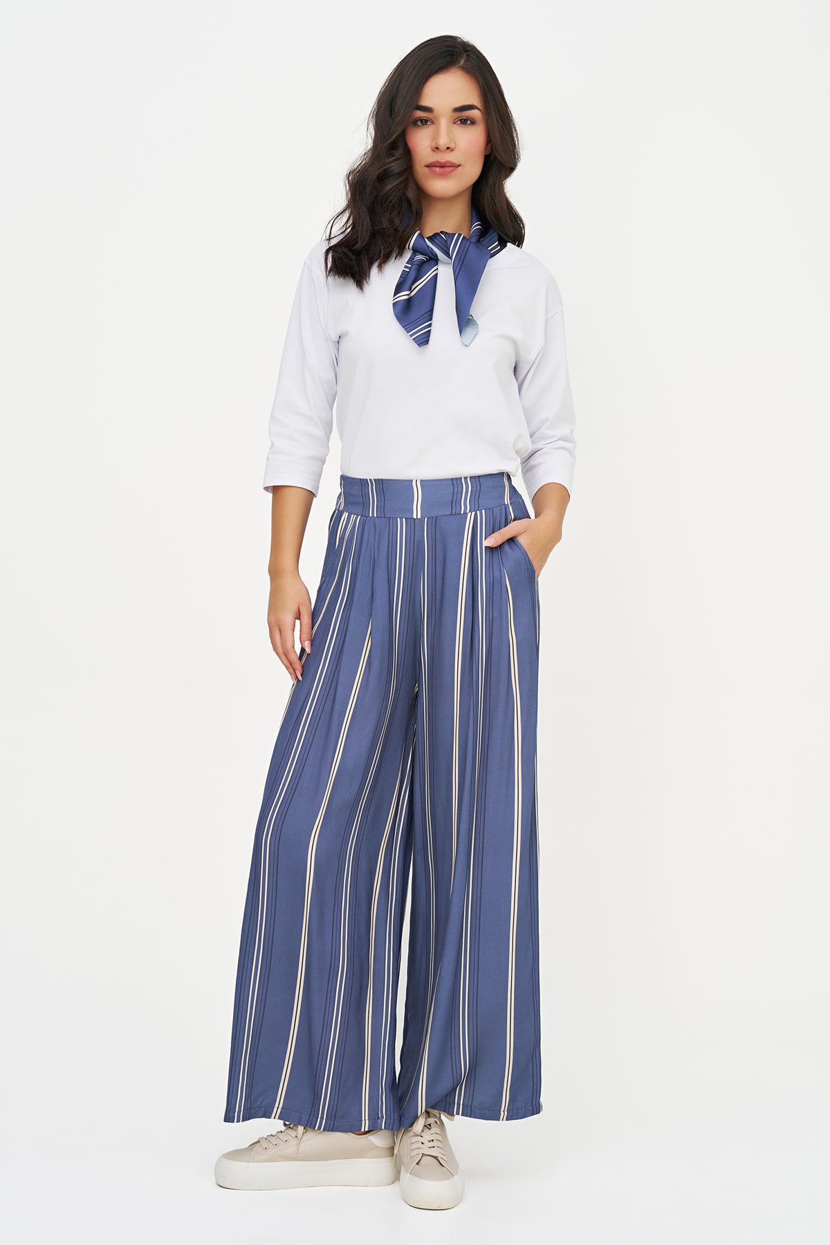 A woman modeling a white long-sleeve blouse with a blue neck scarf and high-waisted, wide-leg blue trousers with white vertical stripes, paired with white sneakers, against a plain white background.
