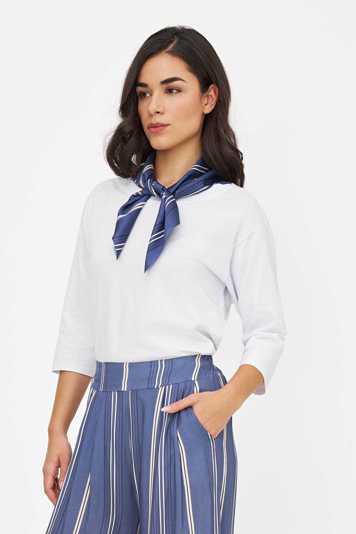 A woman wearing a white three-quarter sleeve top, a navy blue striped neck scarf, and matching navy blue and white vertically striped trousers, photographed against a plain light gray studio background.