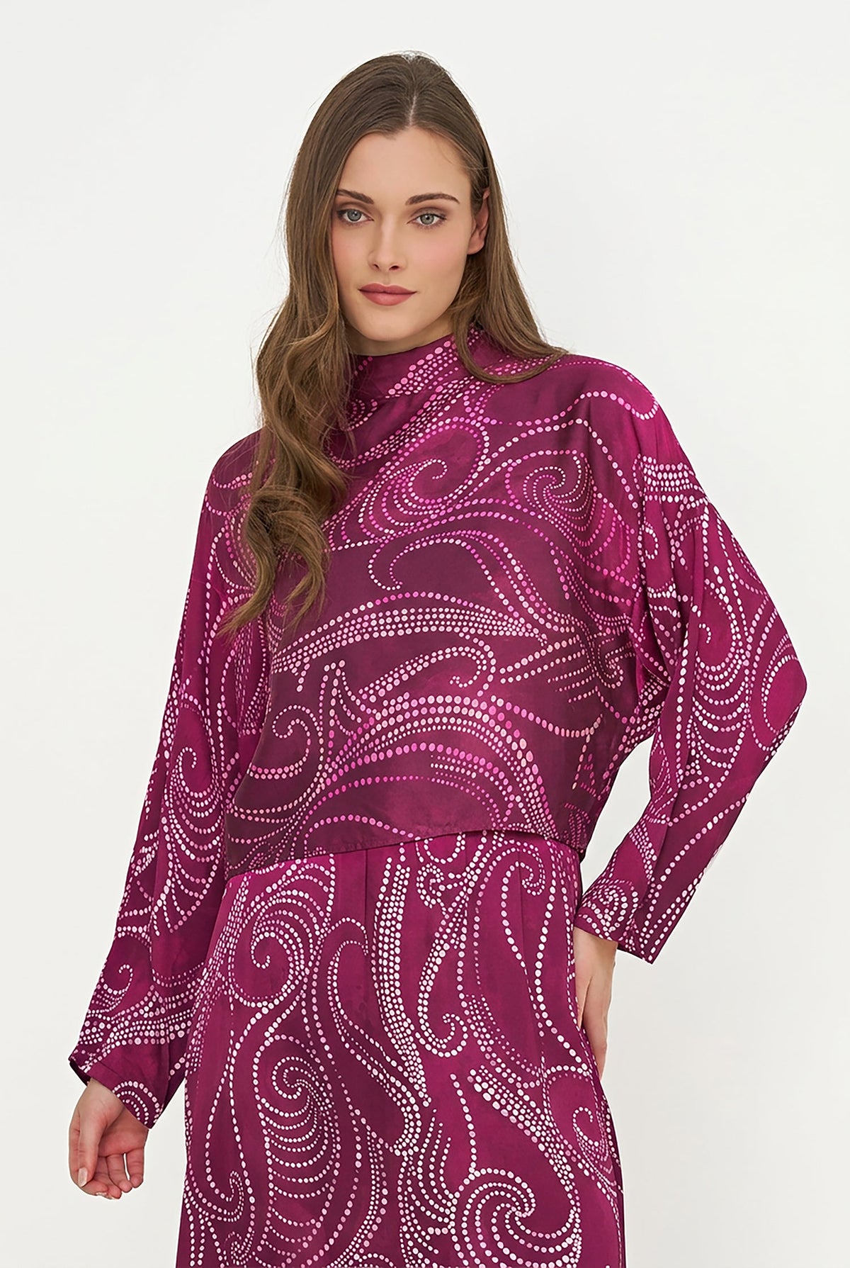 A woman modeling a high-neck, long-sleeve top and matching skirt set in a deep magenta-purple color with a white dotted paisley swirl pattern, set against a plain white studio background.