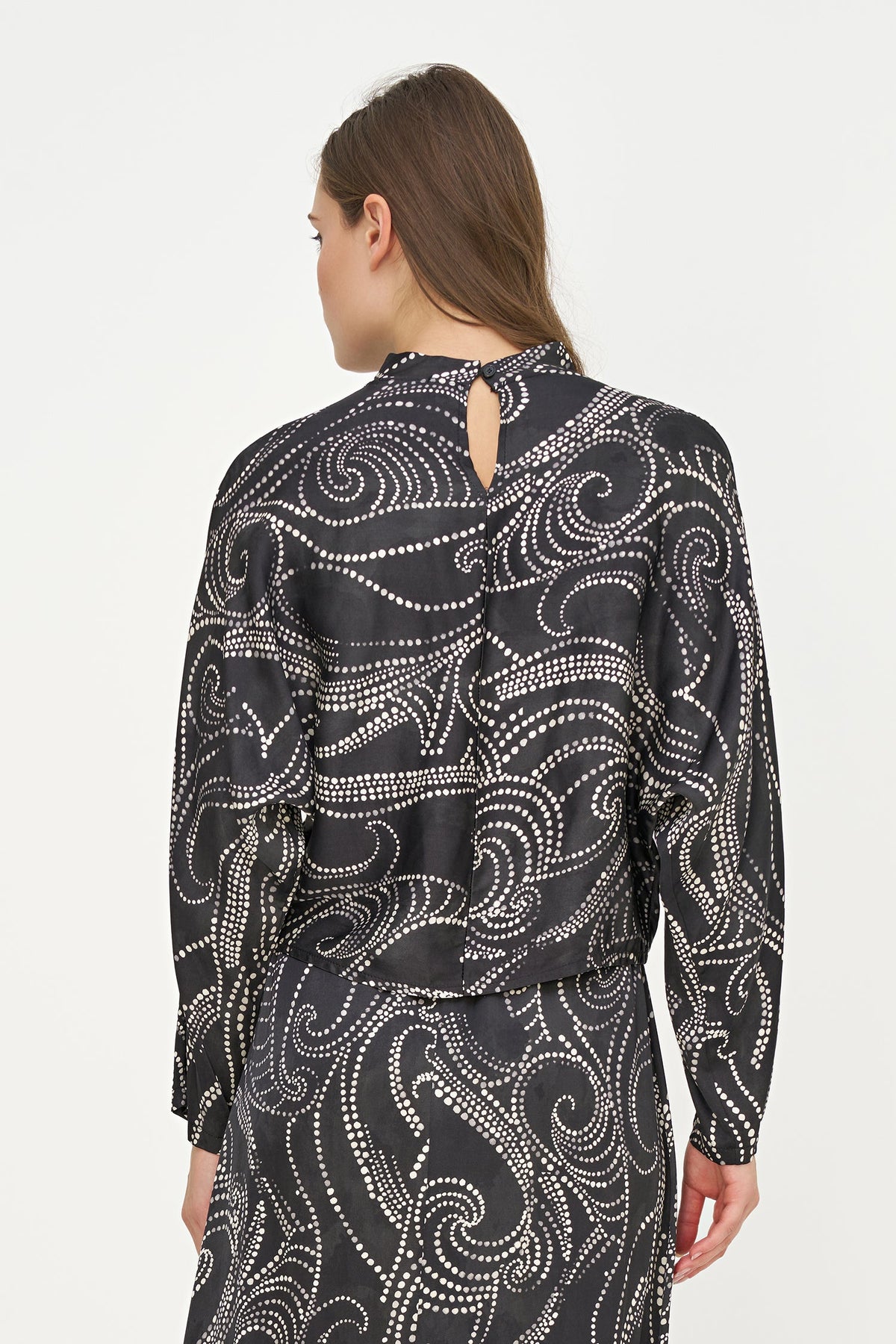 A woman shown from the back wearing a black long-sleeve blouse with a white abstract swirl pattern and a small keyhole cutout at the neckline, against a plain light background.