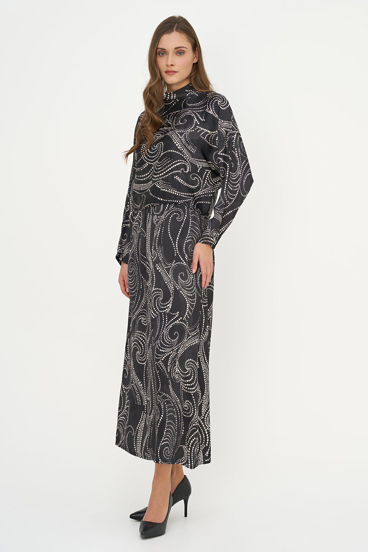 A woman modeling a long-sleeved black midi dress with a white abstract swirl pattern, paired with black high heels, against a plain white studio background.