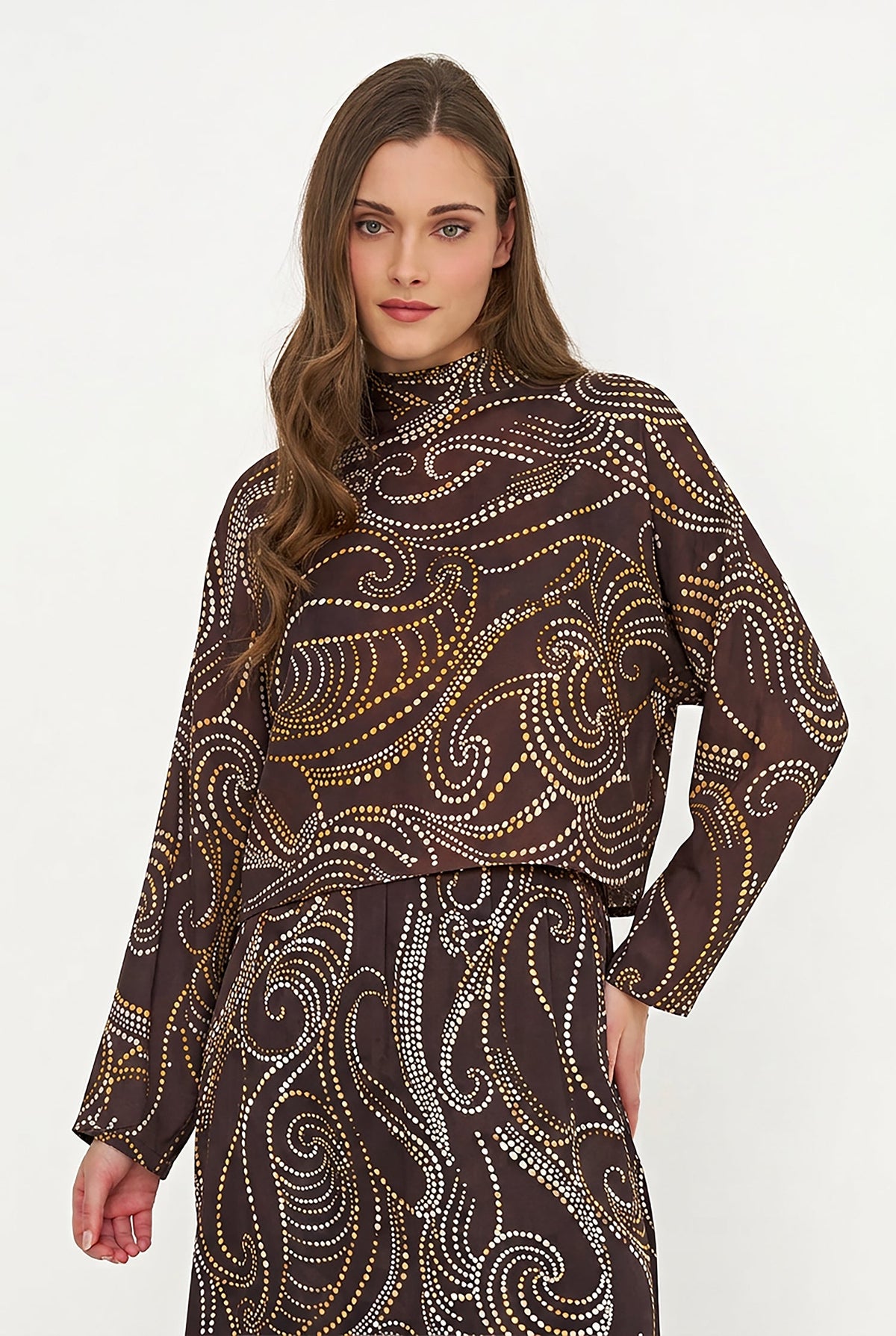 A woman modeling a dark brown long-sleeve mock-neck top with a swirling paisley pattern made of gold and white dotted lines, paired with a matching skirt, against a plain white studio background.