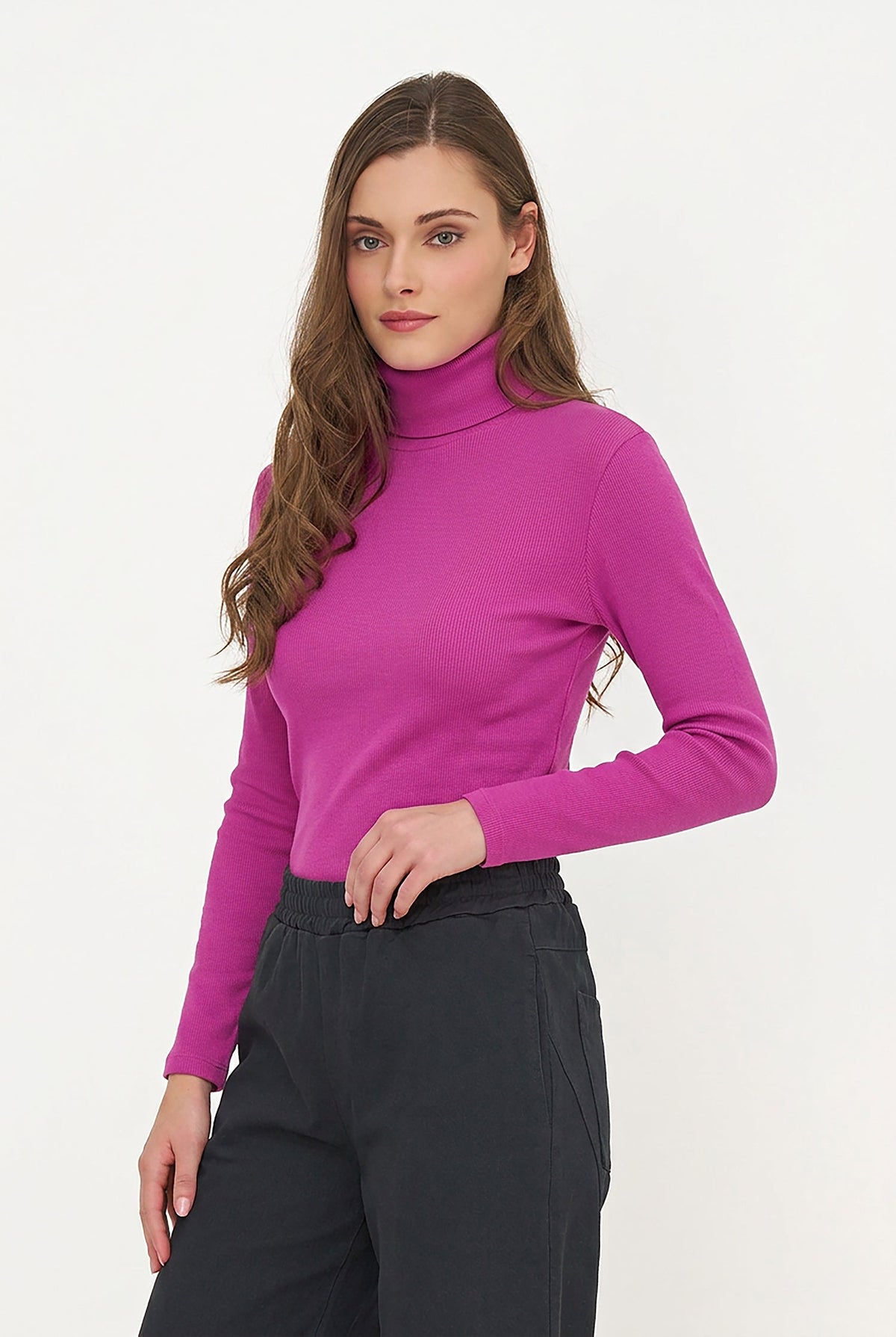 A woman modeling a bright magenta long-sleeve turtleneck sweater paired with dark charcoal trousers, set against a plain white studio background.