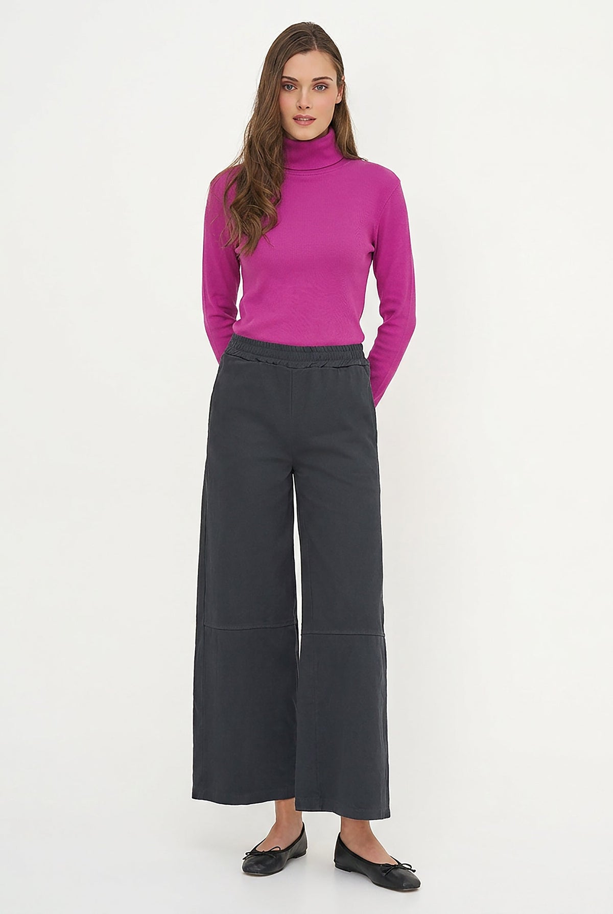 A woman modeling a bright magenta turtleneck sweater paired with dark charcoal wide-leg cropped pants and black flats, standing against a plain white studio background.