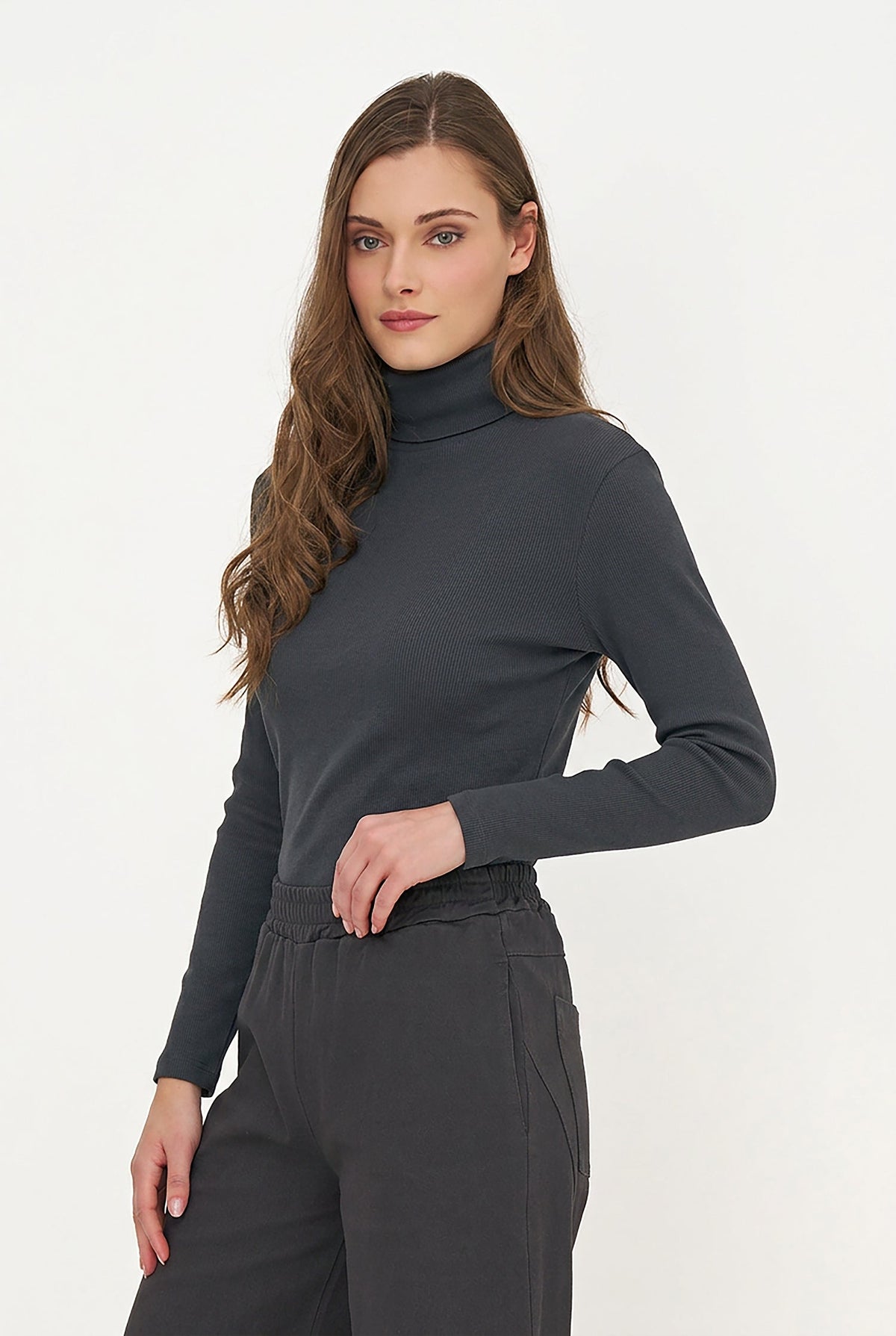 A woman modeling a dark charcoal gray long-sleeve turtleneck top with matching dark gray trousers, photographed against a clean white studio background.