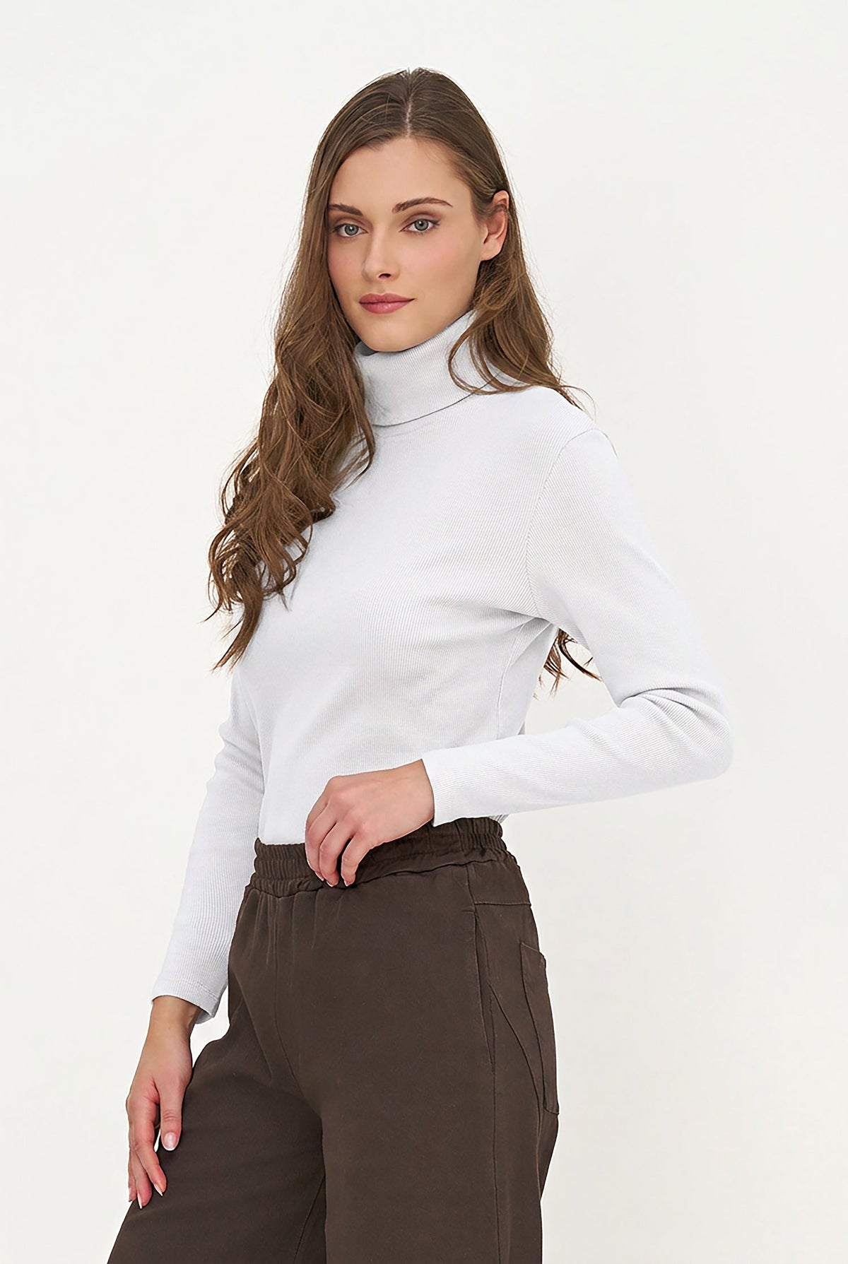 A woman modeling a white long-sleeve turtleneck top paired with dark brown trousers, photographed against a clean white studio background.
