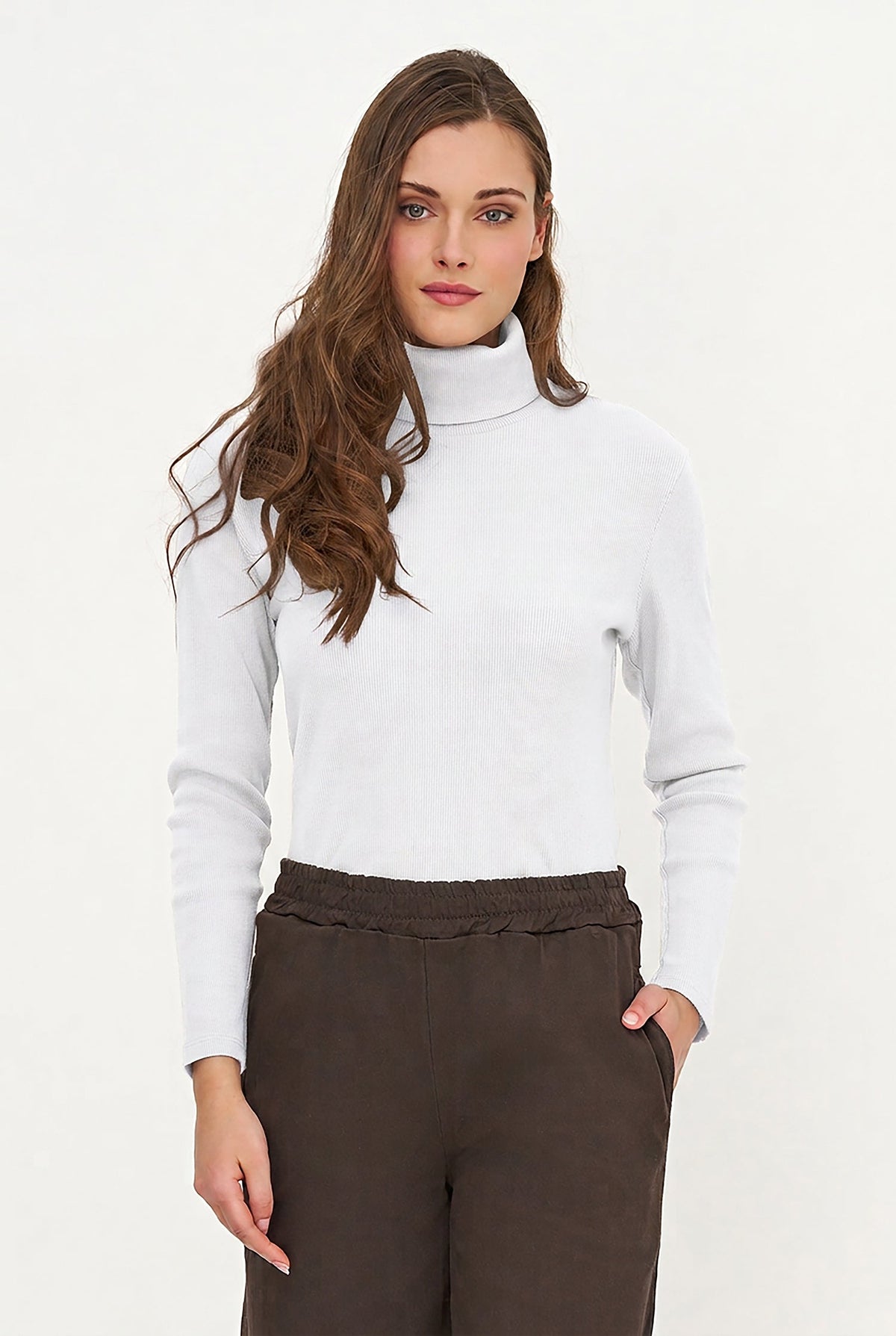 A woman modeling a white ribbed turtleneck sweater paired with dark brown trousers, shot against a clean white studio background.