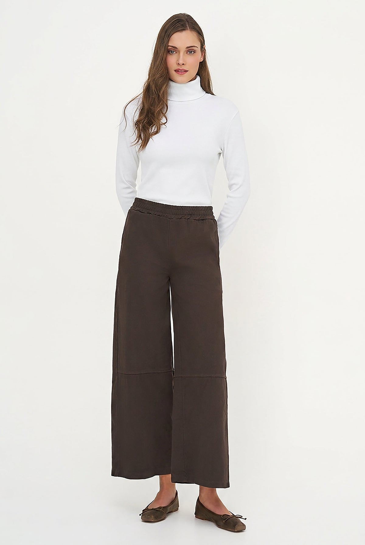 A woman modeling a white long-sleeve mock neck top and dark brown wide-leg cropped pants, paired with brown flat shoes, against a plain white studio background.
