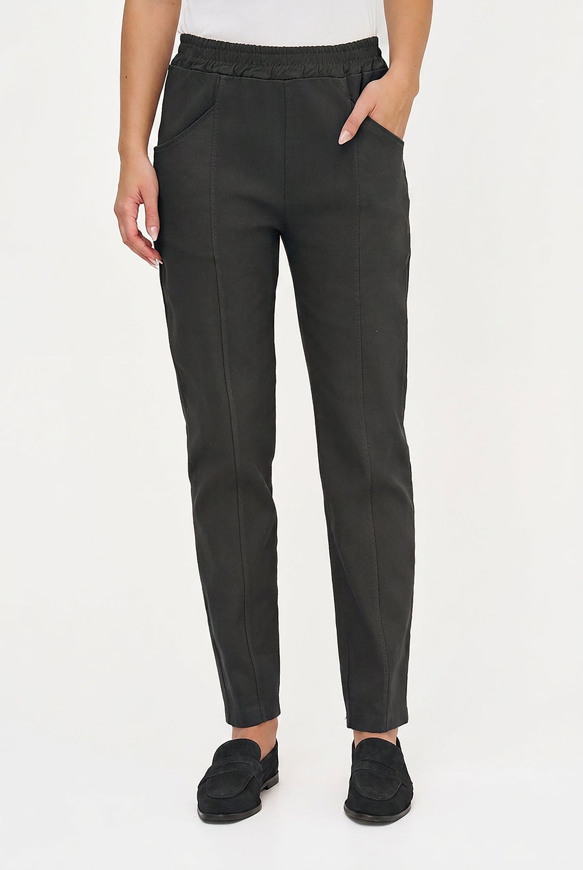 A person wearing dark charcoal slim-fit trousers with an elastic waistband, side pockets, and a straight tapered leg, paired with black slip-on loafers against a plain white studio background.