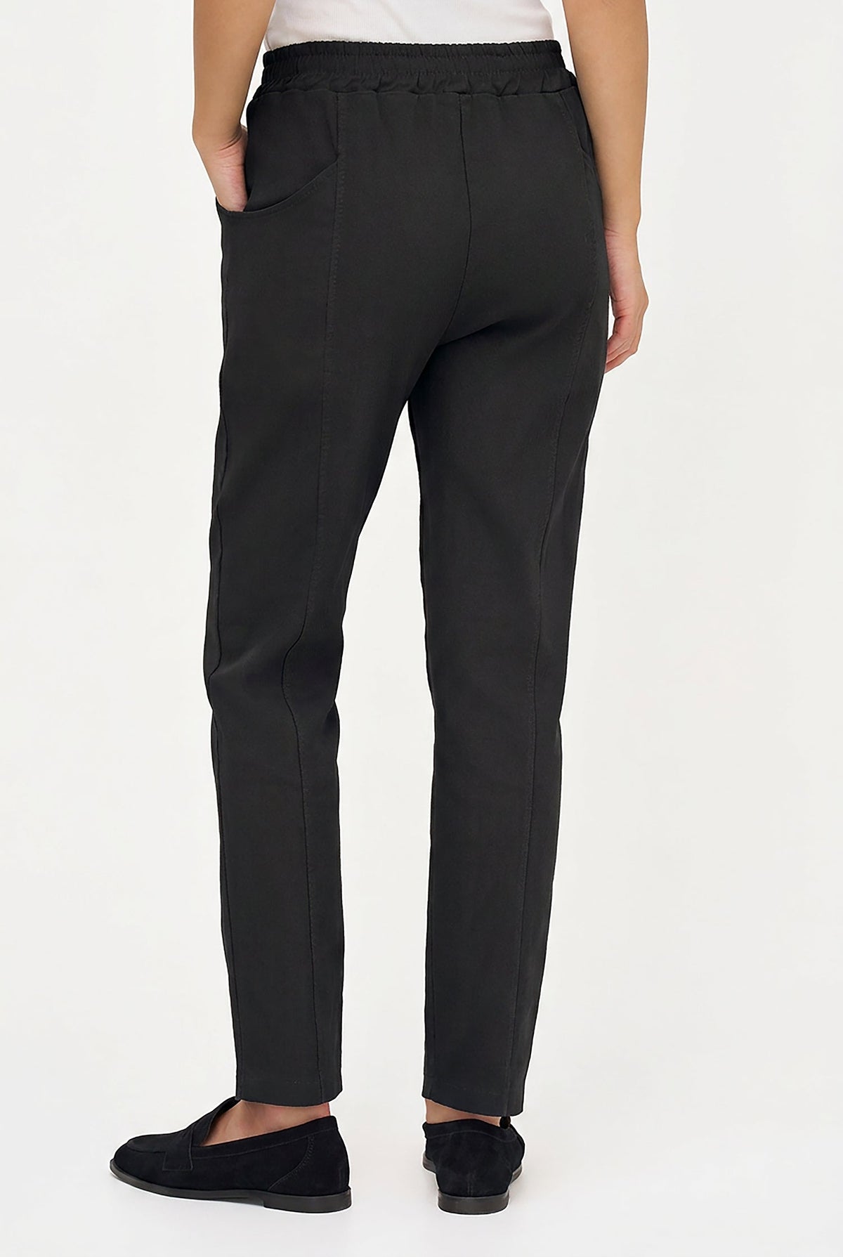 A person shown from the back wearing slim black trousers with side pockets and black slip-on shoes against a plain light background.