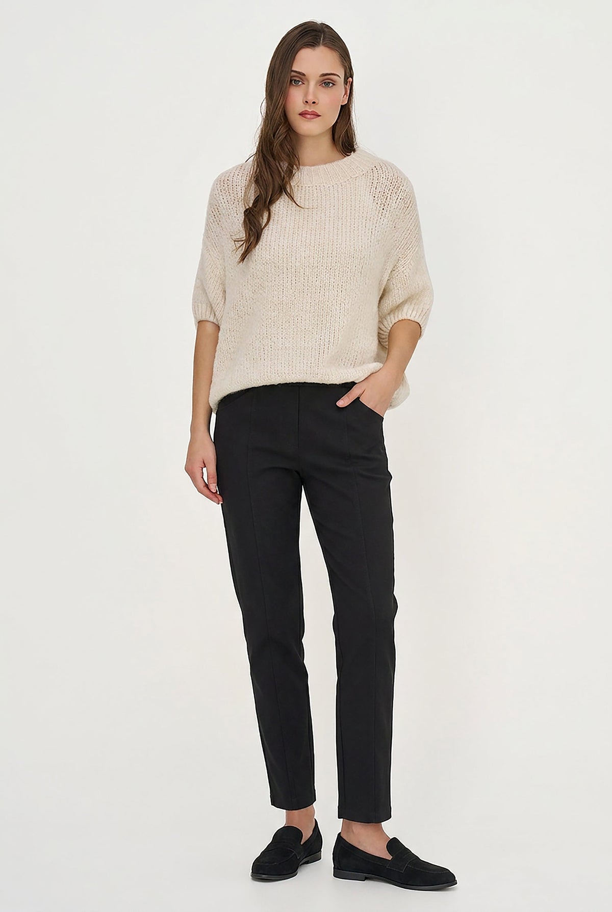 A woman modeling a cream-colored textured knit sweater and black slim-fit trousers with black slip-on shoes, standing against a plain white studio background.