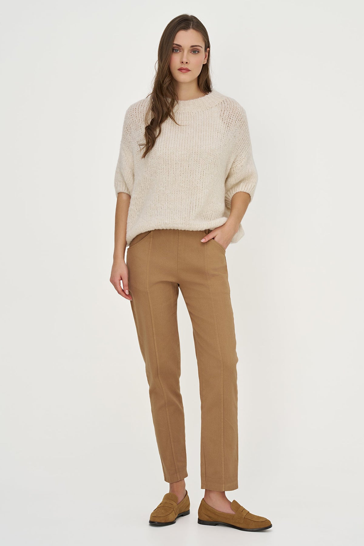 A woman modeling a cream-colored chunky knit sweater and tan trousers against a plain white studio background.