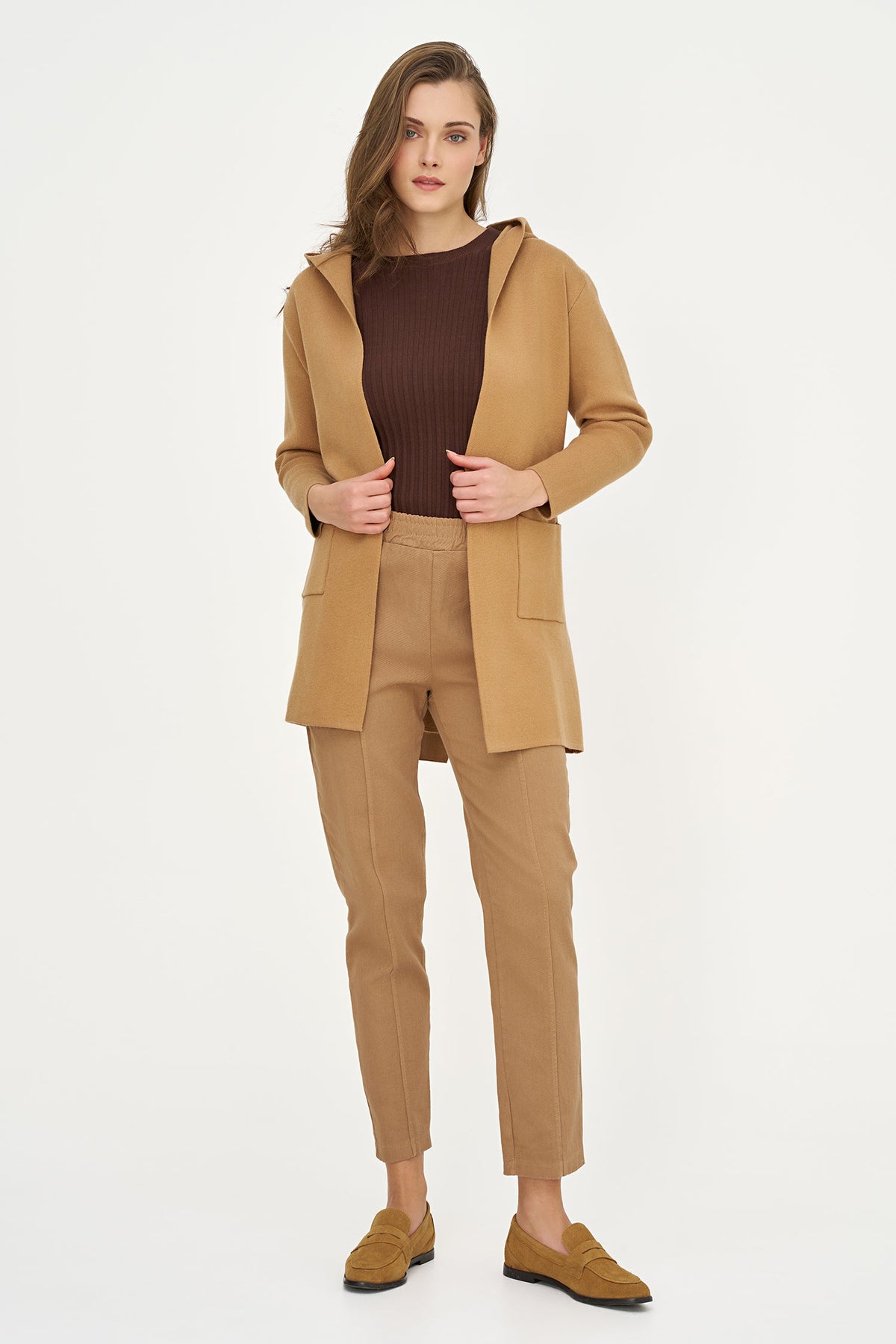 A woman modeling a camel-colored hooded jacket, matching trousers, a dark brown ribbed top, and brown suede loafers against a plain white studio background.