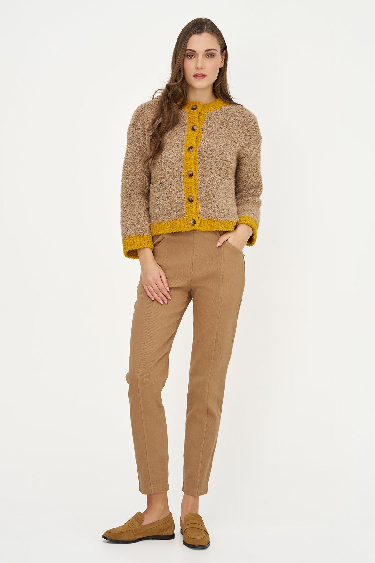 A woman modeling a cropped, textured knit cardigan in beige with a mustard yellow button placket, collar, cuffs, and hem, paired with tan trousers and brown suede loafers, against a plain white studio background.