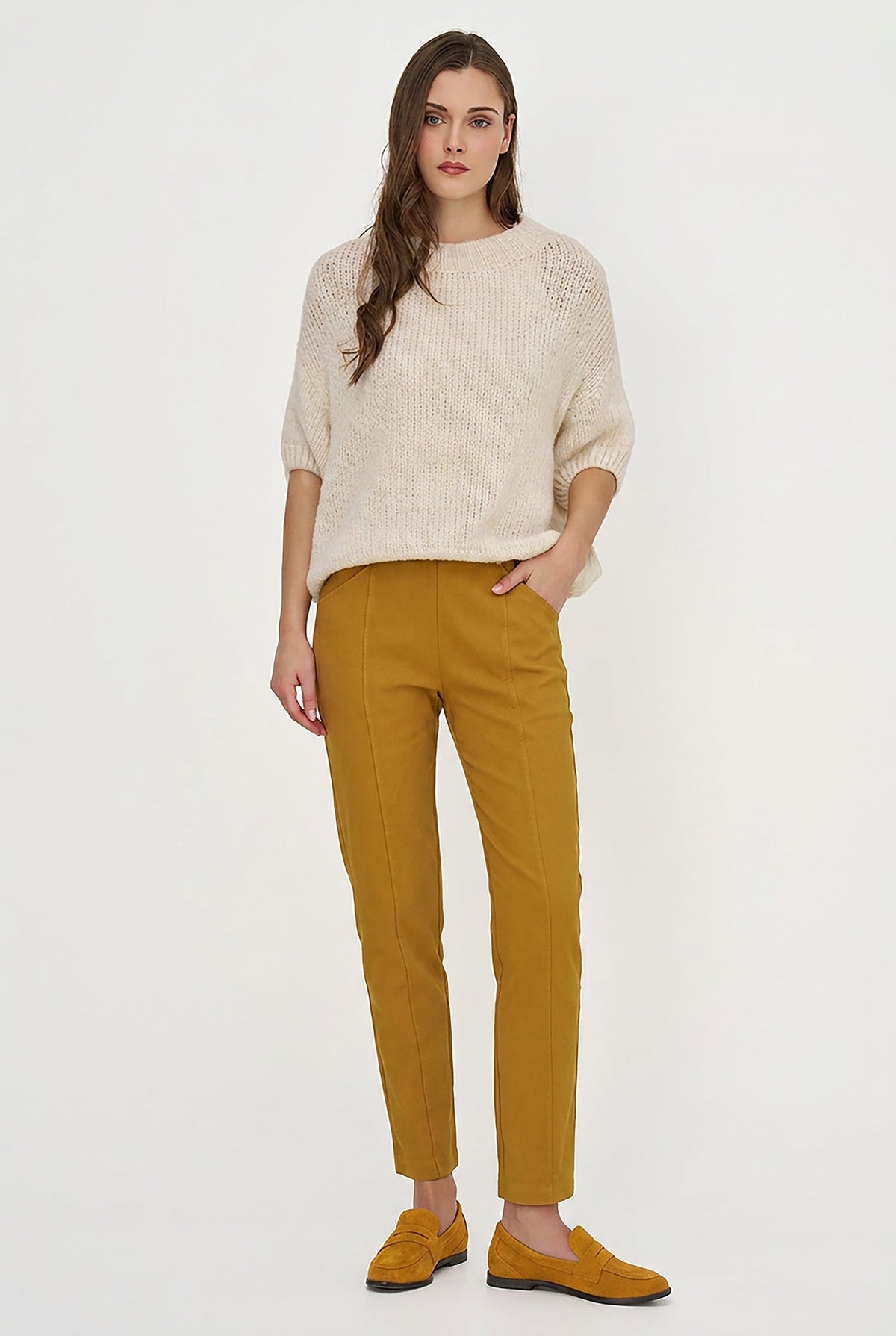 A woman modeling a cream-colored textured knit sweater and mustard yellow trousers with matching suede loafers, standing against a plain white studio background.