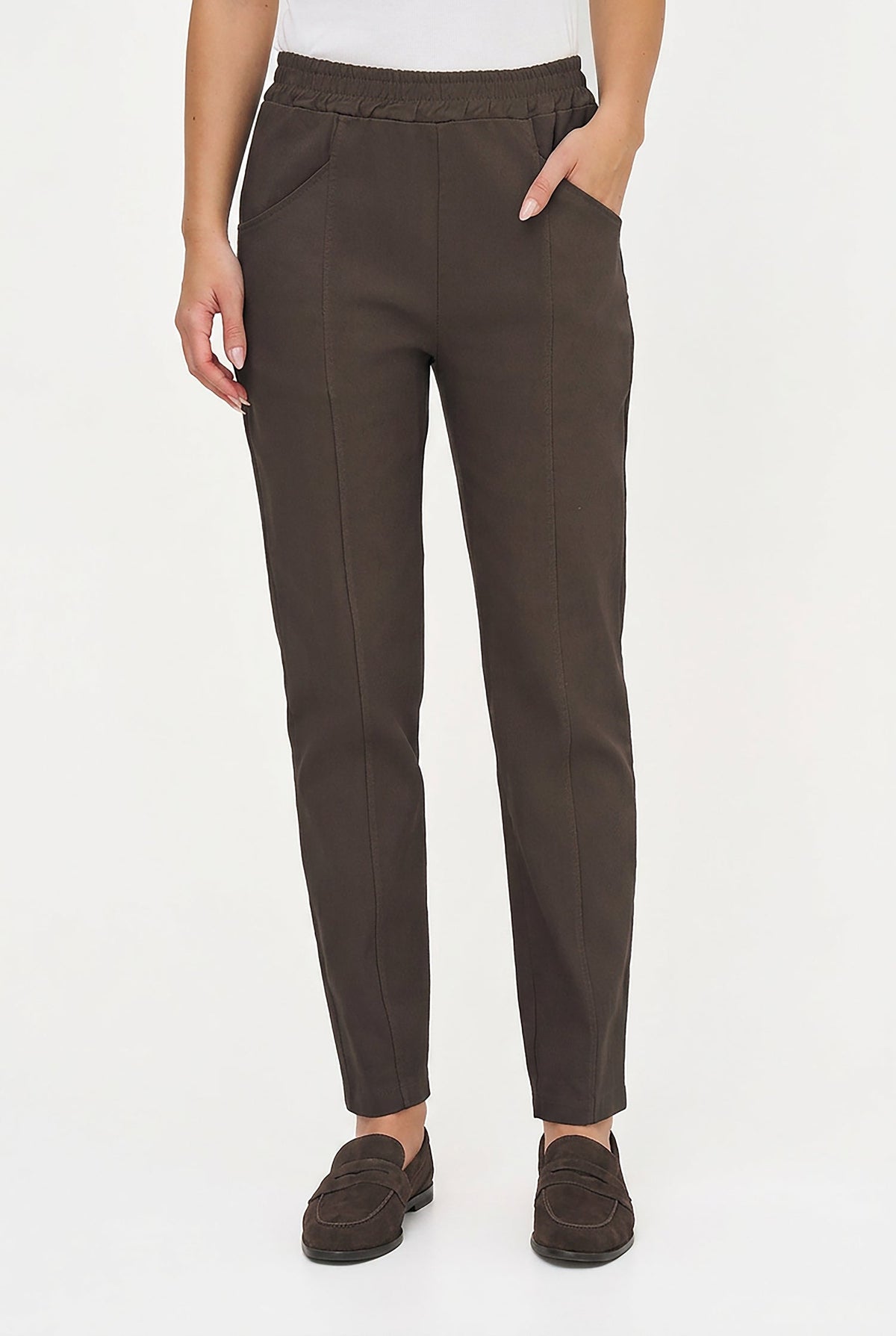 A person wearing dark brown slim-fit trousers with side pockets, paired with a white top and dark brown loafers, shown from the waist down against a plain white studio background.