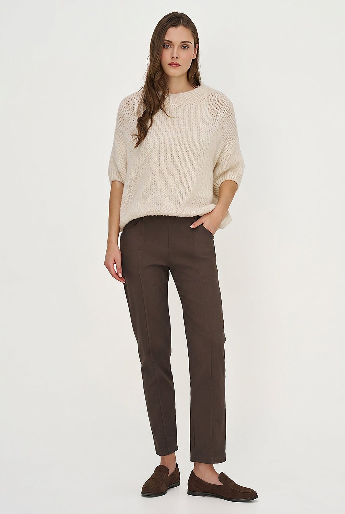A woman modeling a cream-colored textured knit sweater and dark brown slim-fit trousers with matching brown suede loafers, set against a plain white studio background.