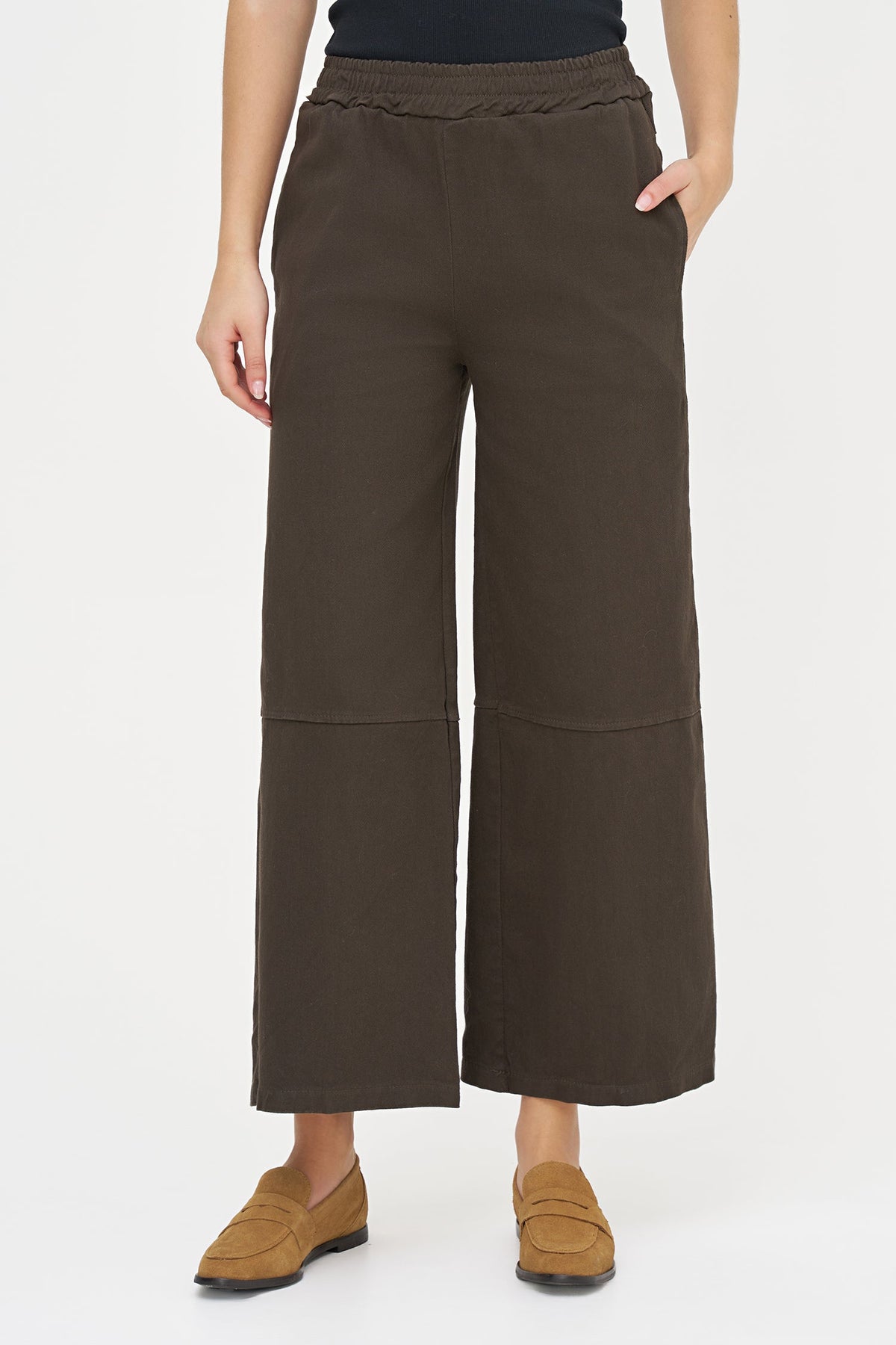 A model wearing dark brown wide-leg cropped pants with an elastic waistband and side pockets, paired with tan loafers, against a plain white studio background.