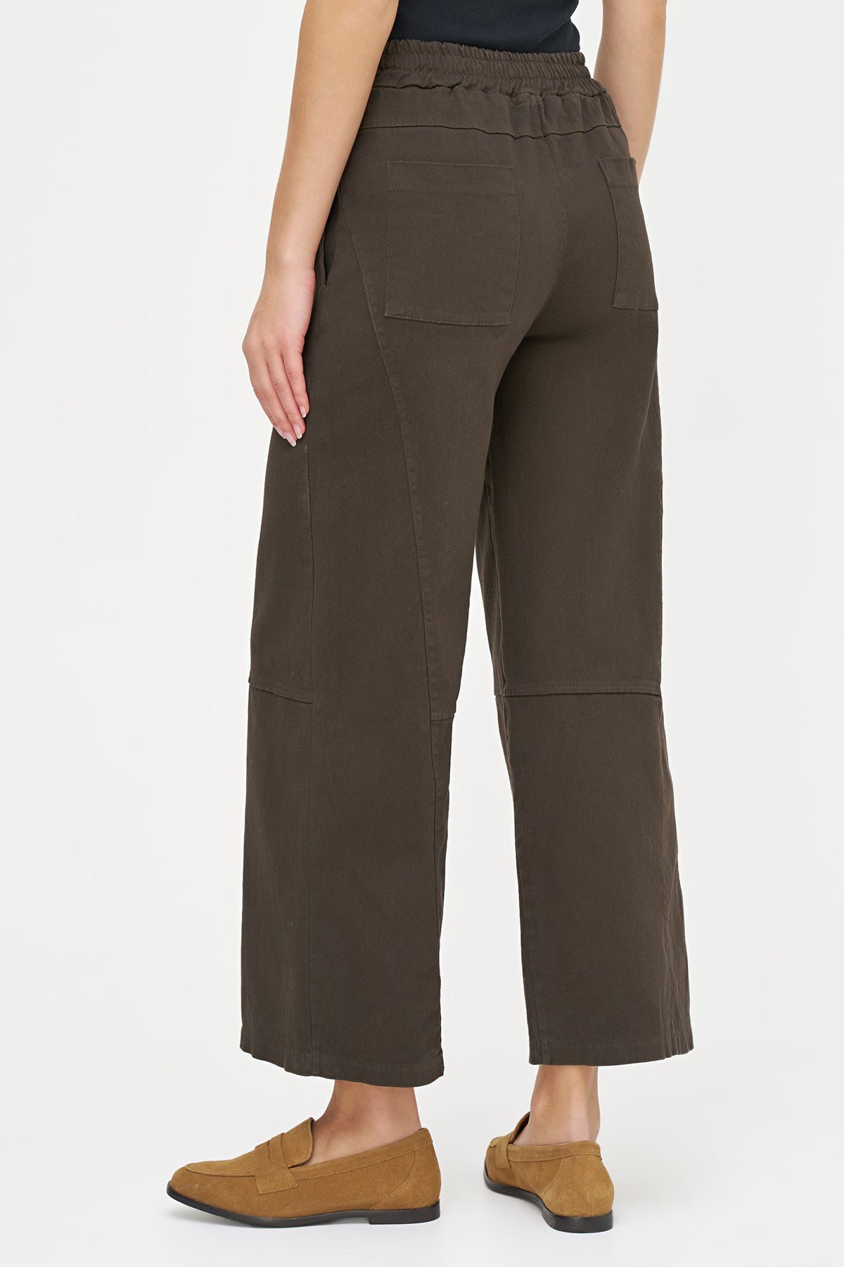 A person shown from the back wearing dark brown cropped wide-leg pants with side pockets and tan slip-on loafers, photographed against a clean light gray studio background.