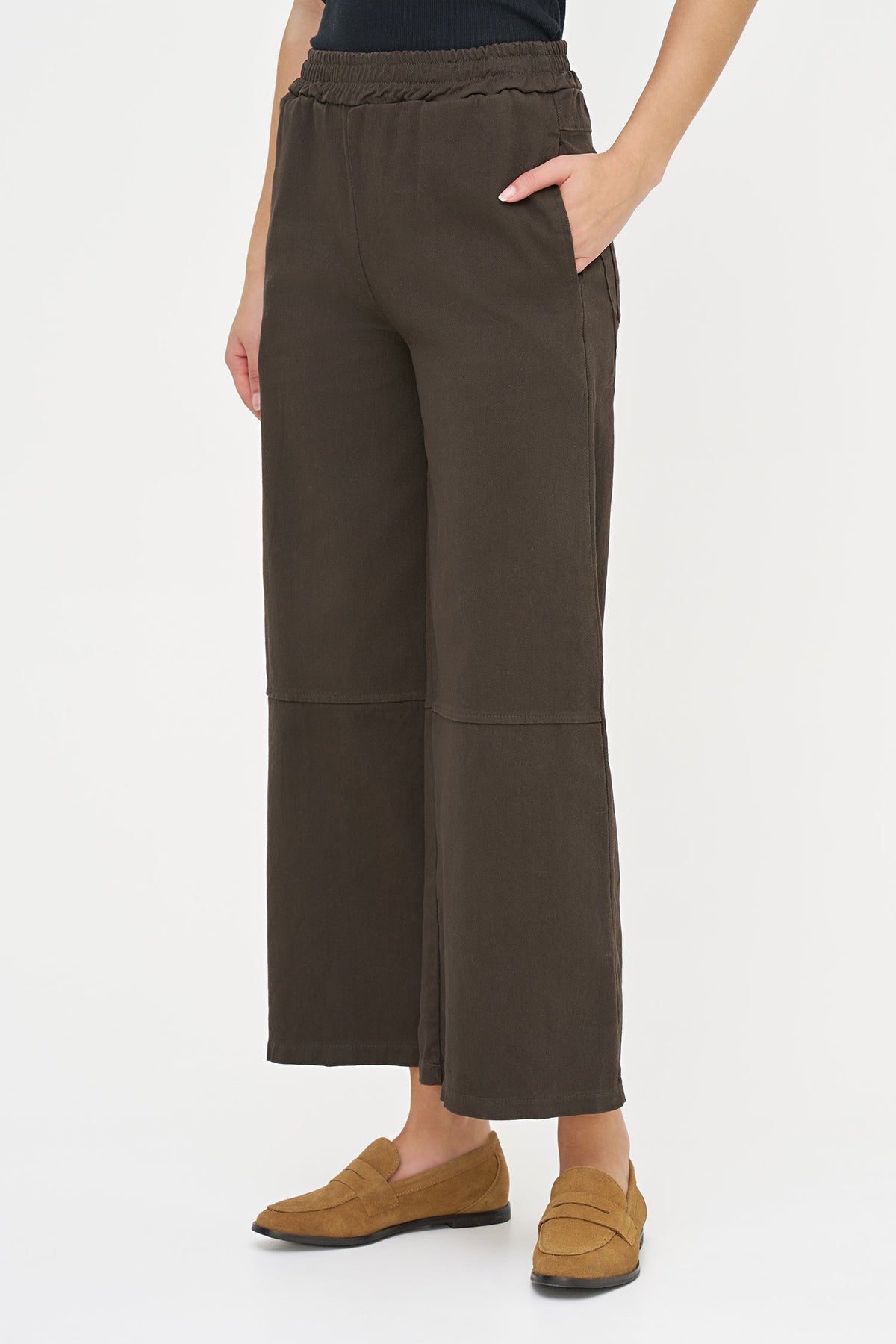 A model wearing dark brown wide-leg cropped pants with side pockets and a gathered elastic waistband, paired with tan loafers against a plain white studio background.