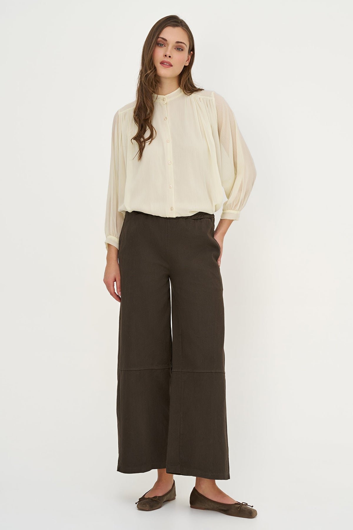 A woman modeling a cream-colored long-sleeve blouse with voluminous sleeves and dark brown wide-leg cropped pants, paired with brown flats, against a plain white studio background.