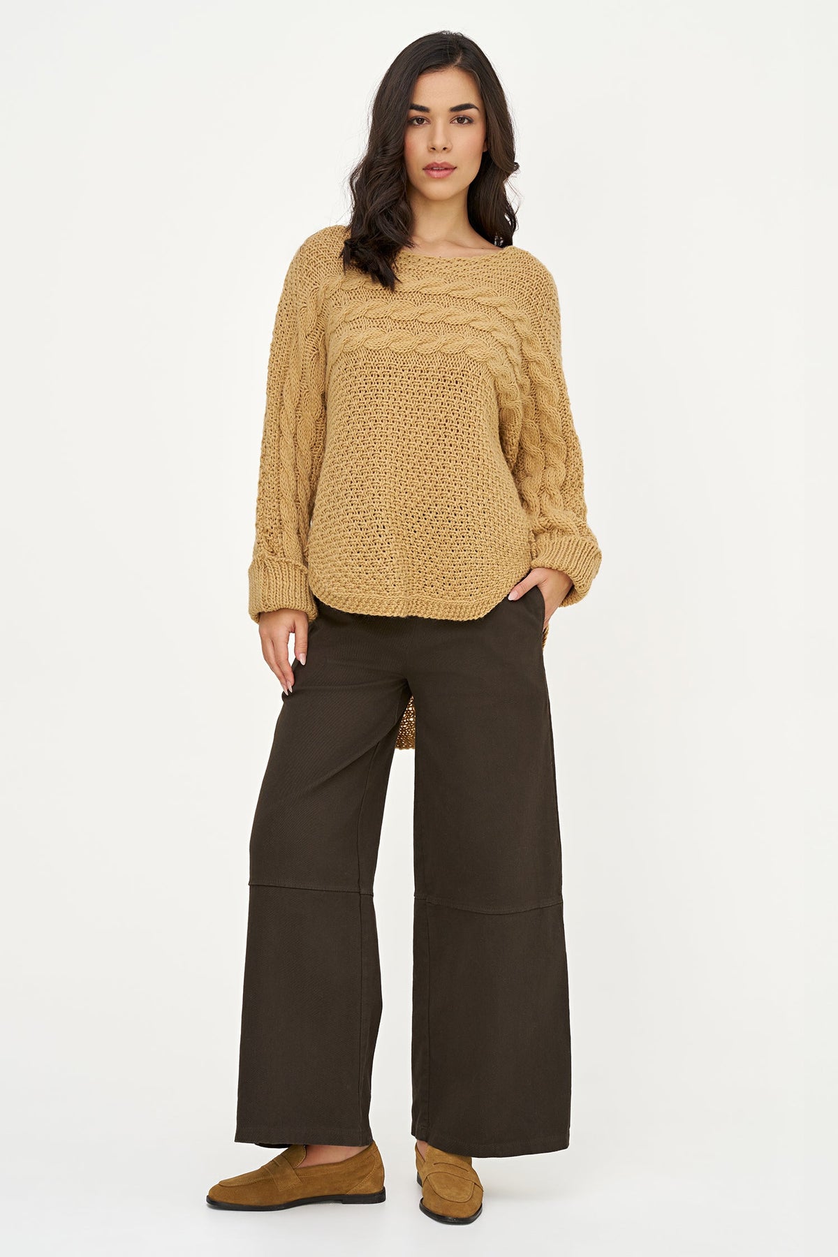 A woman modeling a chunky cable-knit sweater in camel beige and wide-leg dark brown pants, standing against a plain white studio background.