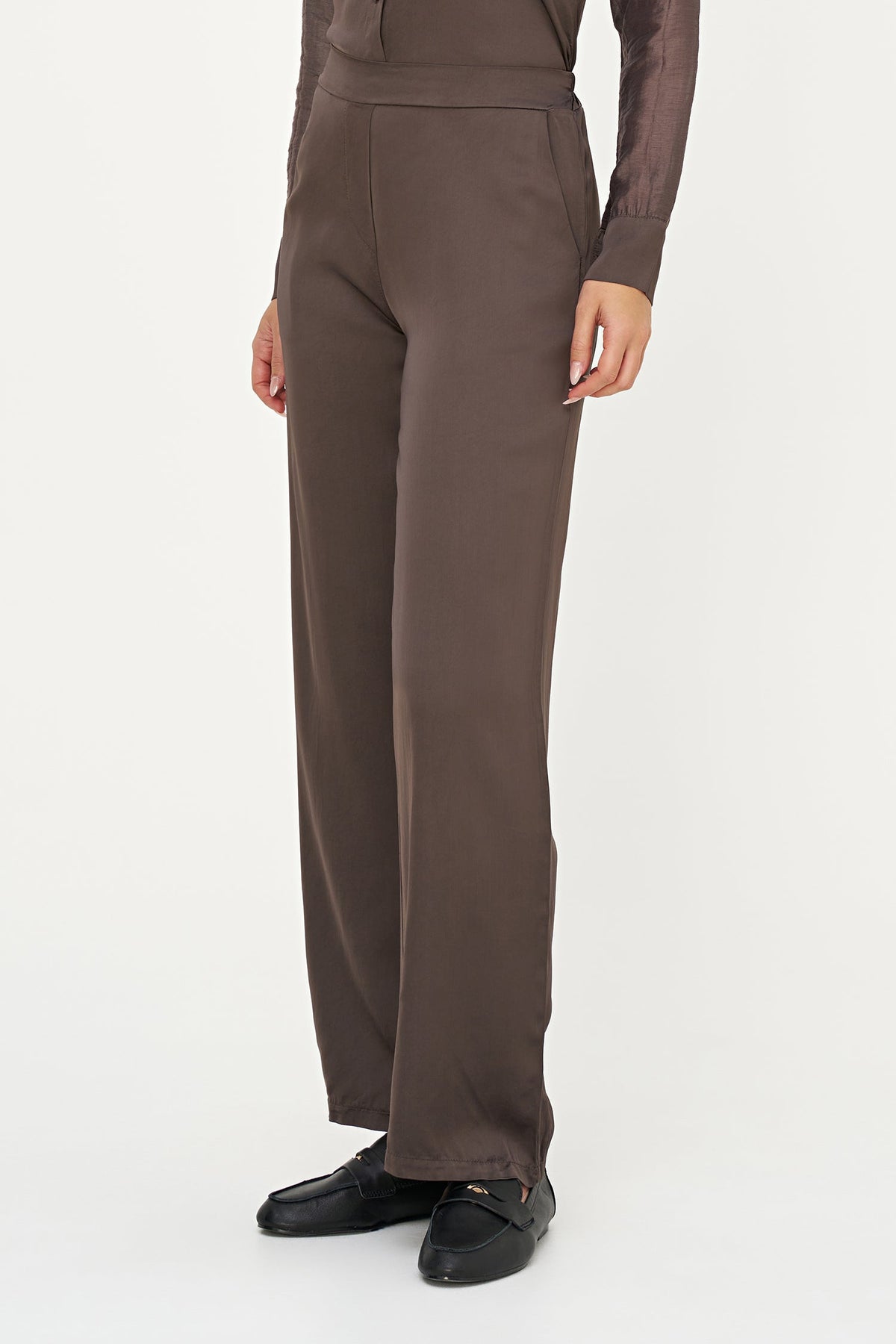 A model wearing high-waisted, wide-leg brown trousers paired with a matching brown sheer long-sleeve top and black loafers, shown from the waist down against a plain white studio background.