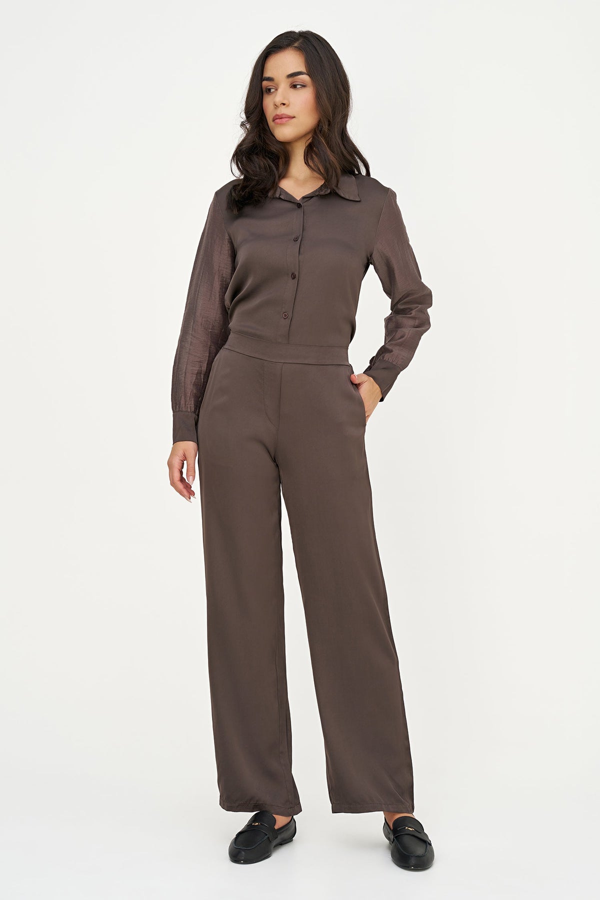 A woman modeling a matching two-piece outfit consisting of a long-sleeve button-up shirt and wide-leg trousers in a dark brown color, paired with black loafers, against a plain white studio background.