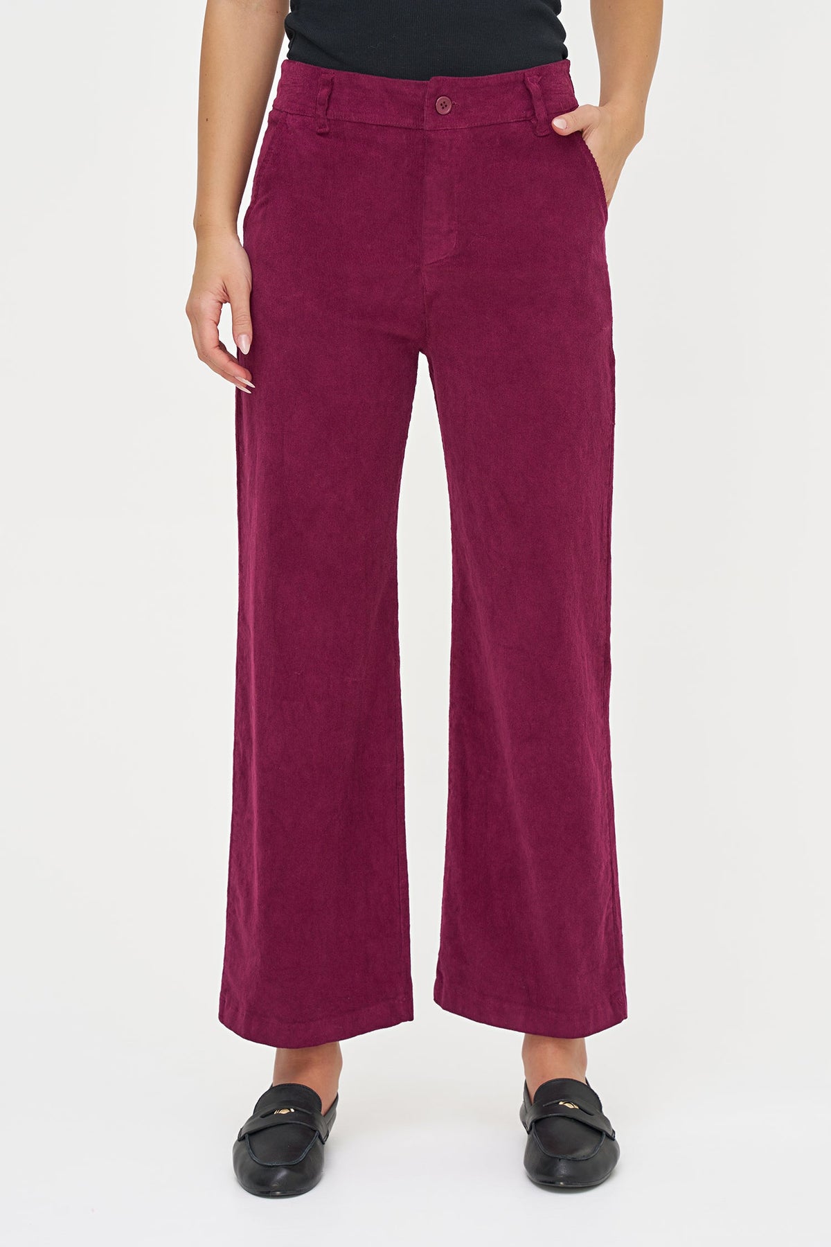 A model wearing a pair of wide-leg cropped pants in a deep magenta corduroy fabric, styled with a black top and black loafers against a plain white studio background.