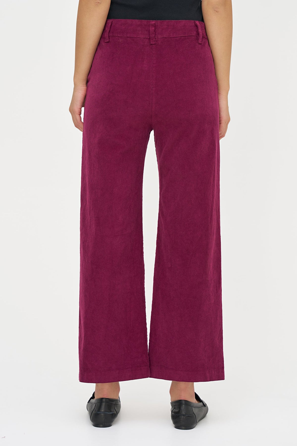 A person shown from the waist down wearing high-waisted, wide-leg trousers in a deep magenta-burgundy corduroy fabric, paired with black flats, against a plain white studio background.