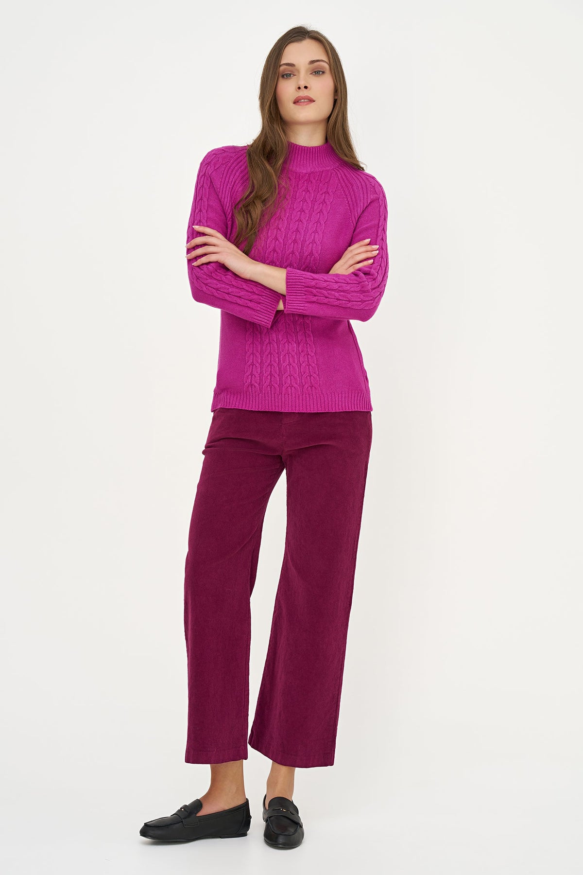 A woman modeling a bright magenta cable-knit mock-neck sweater paired with matching magenta wide-leg trousers and black loafers, set against a plain white studio background.