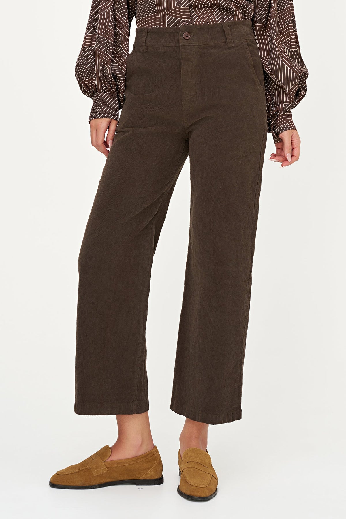 A model wearing dark brown cropped wide-leg trousers, a brown and beige patterned blouse, and tan suede loafers, shown from the waist down against a plain white studio background.