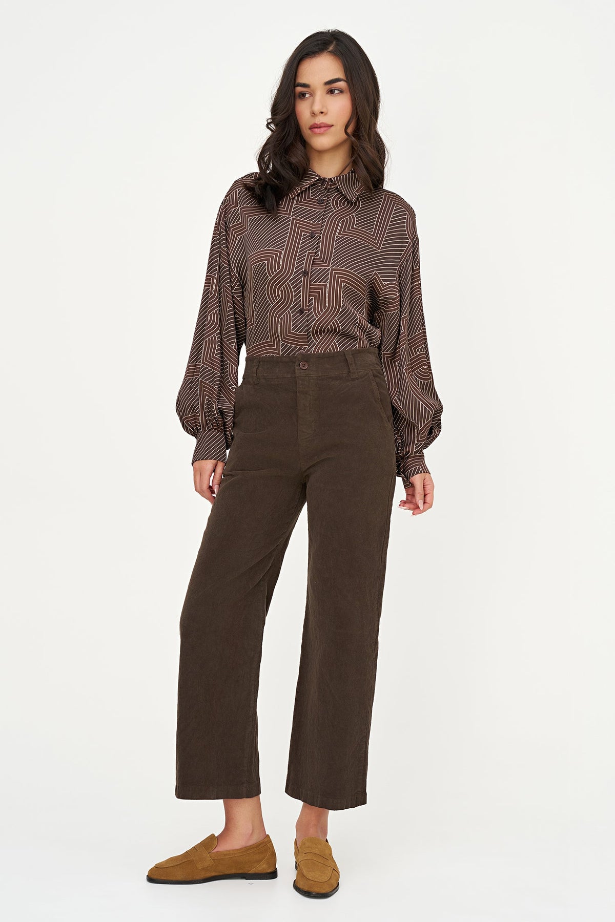 A woman modeling a brown patterned long-sleeve button-up shirt and dark brown cropped trousers, paired with brown suede loafers, against a plain white studio background.