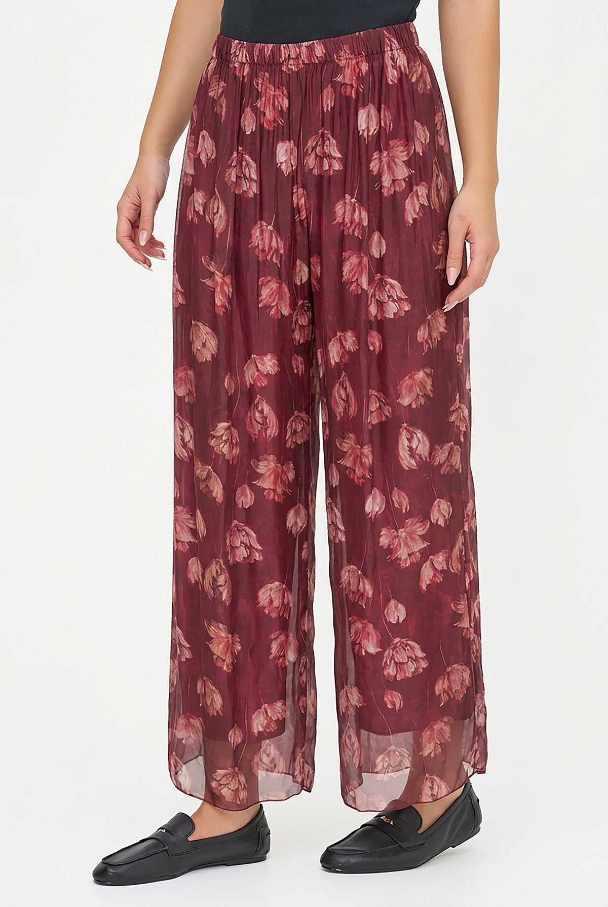 A person wearing loose, wide-leg pleated trousers in a burgundy base with a muted floral print in dusty pink and mauve, paired with black loafers against a plain white studio background.