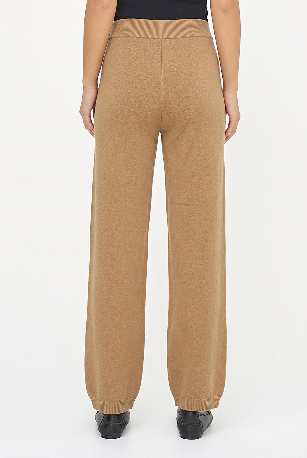 A person shown from the back wearing tan wide-leg knit pants, a black top, and black shoes against a plain white studio background.