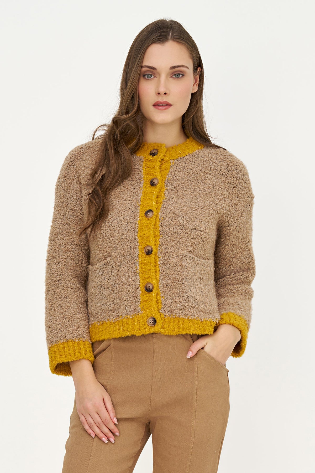 A woman modeling a cropped, textured knit cardigan in a warm beige/tan color with a contrasting mustard yellow button placket, collar, cuffs, and hem, paired with tan trousers against a plain white studio background.