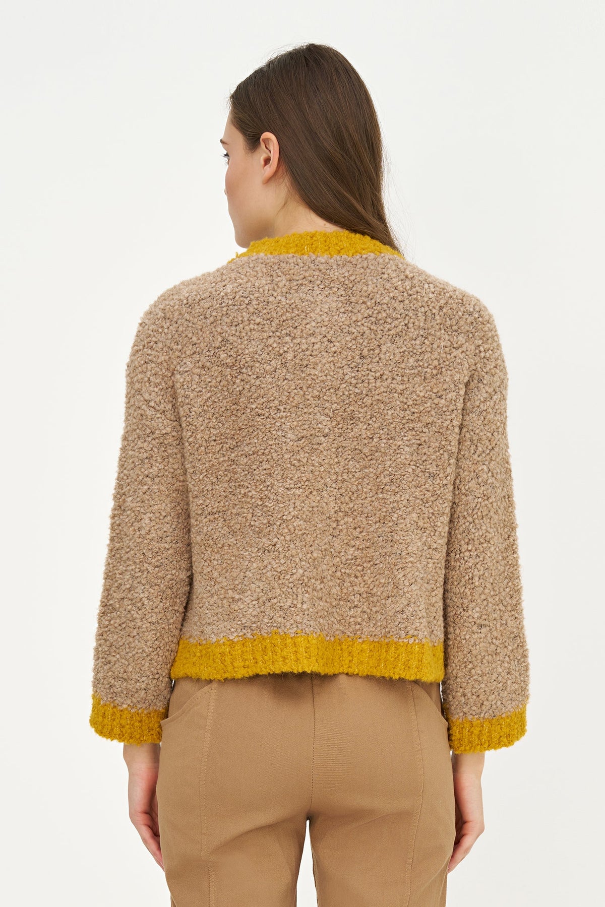 A woman shown from the back wearing a cropped, textured knit cardigan in a heathered beige/tan color with contrasting mustard yellow trim at the collar, hem, and cuffs, paired with tan trousers against a plain white studio background.
