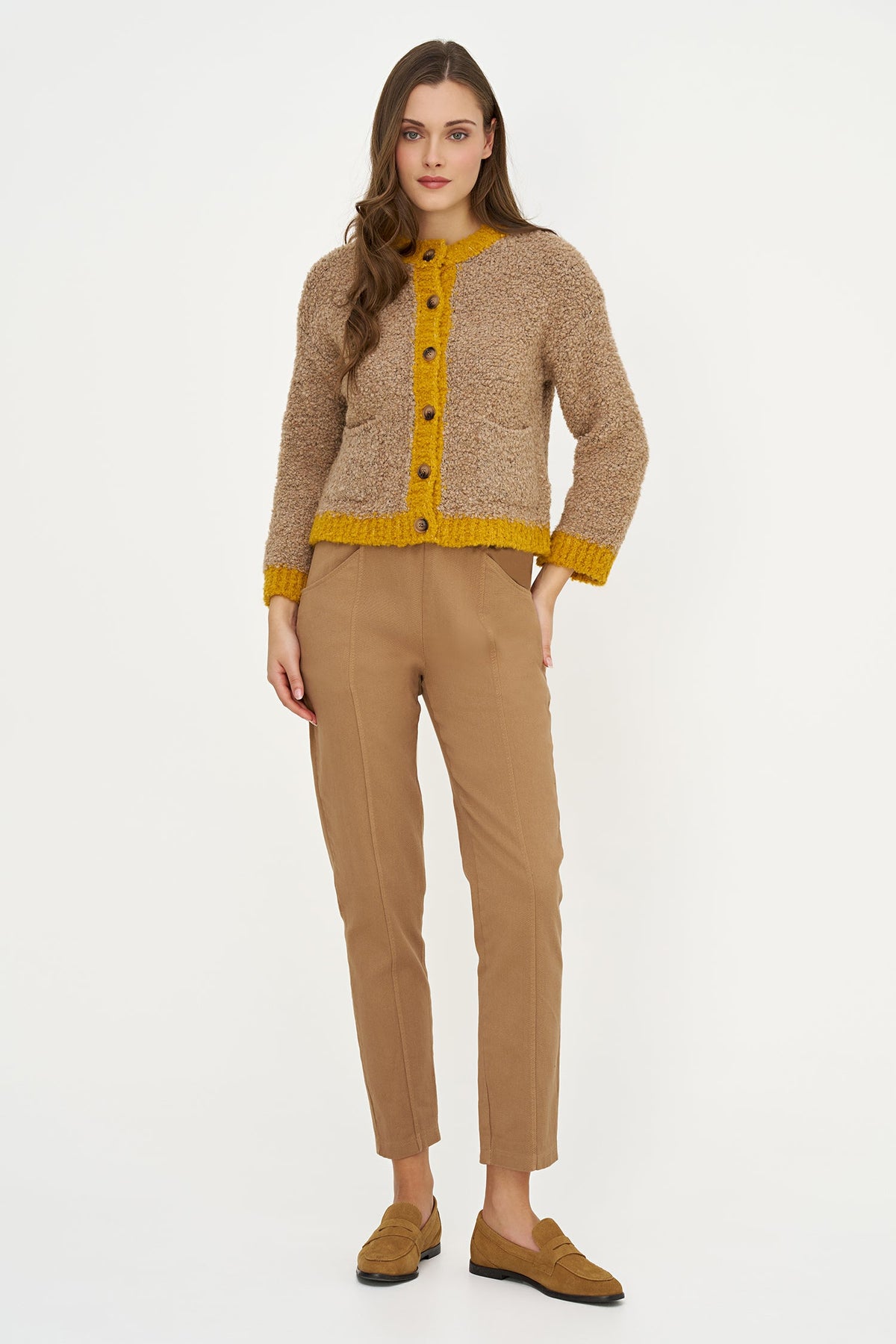 A woman modeling a beige fuzzy knit cardigan with mustard yellow trim and buttons, paired with tan trousers and brown loafers, against a plain white studio background.
