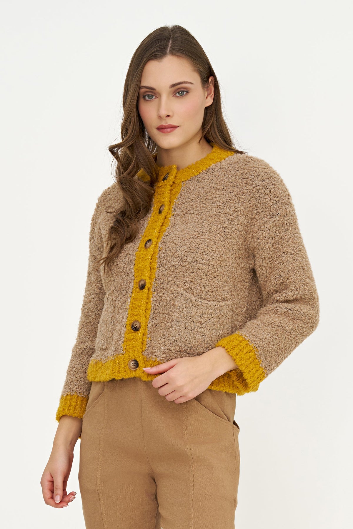 A woman modeling a cropped, textured bouclé cardigan in beige with mustard yellow trim and buttons, paired with tan trousers, against a plain white studio background.