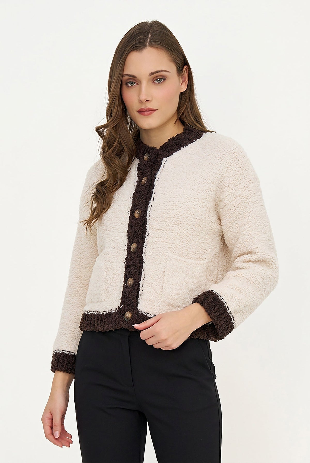 A woman modeling a cropped, textured cream bouclé jacket with dark brown trim, button-front closure, and front pockets, paired with black pants against a plain white studio background.