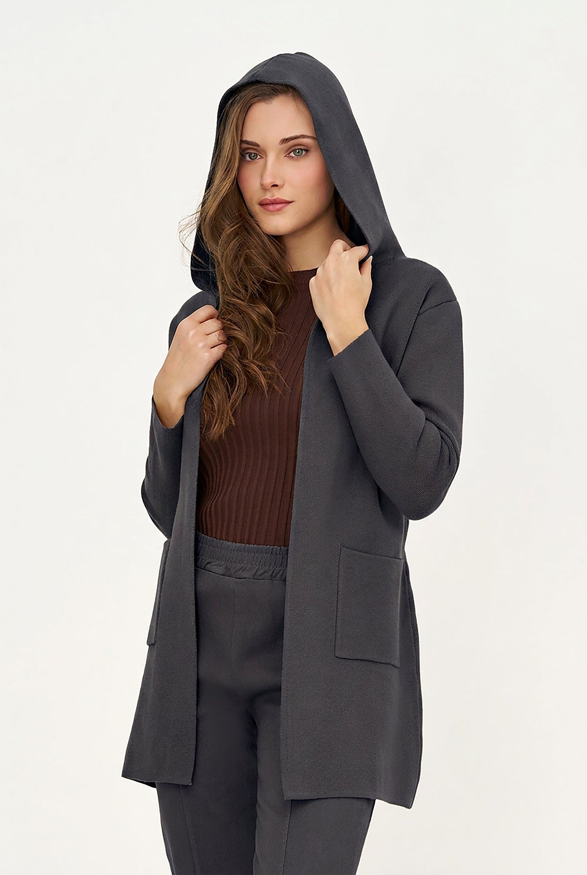 A woman wearing a dark gray hooded cardigan over a brown ribbed top and matching gray pants, posed against a plain light gray studio background.