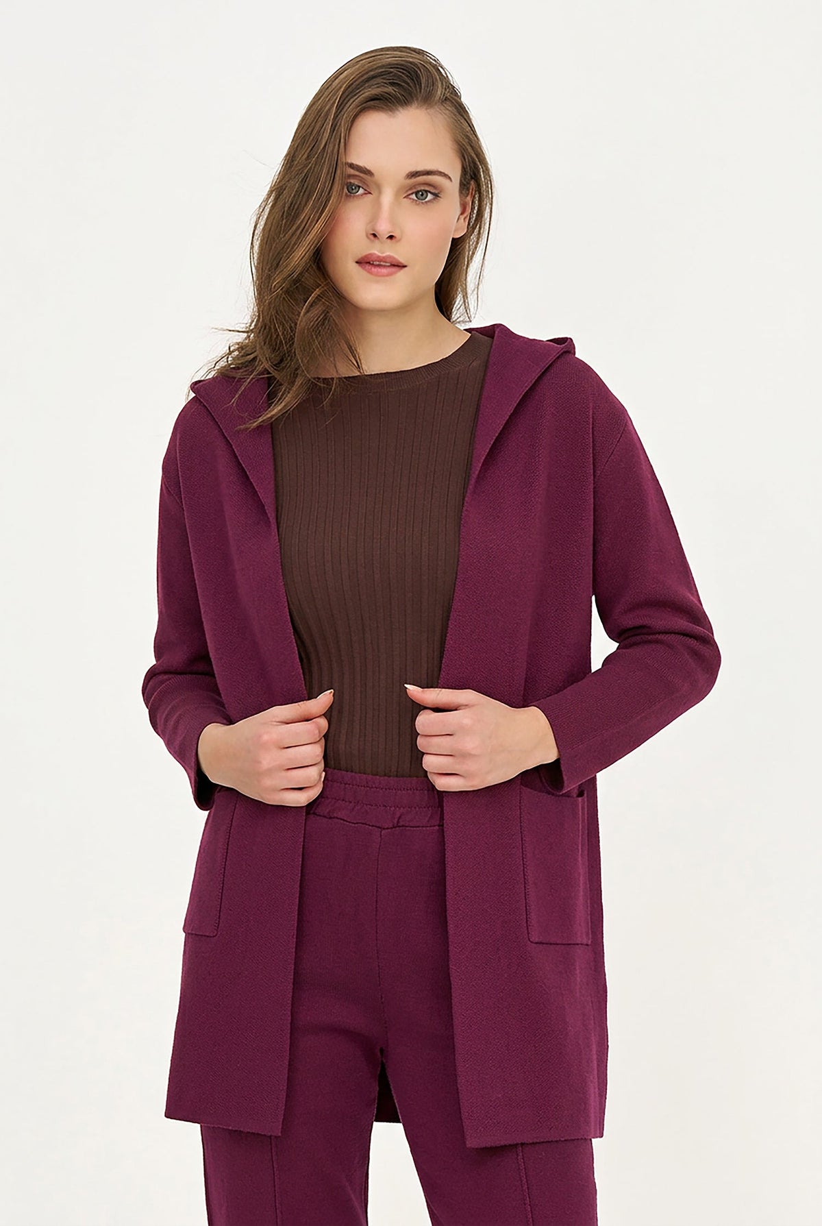 A woman modeling a matching three-piece outfit consisting of a deep plum purple hooded cardigan, a dark brown ribbed knit top, and matching purple trousers, shot against a plain white studio background.