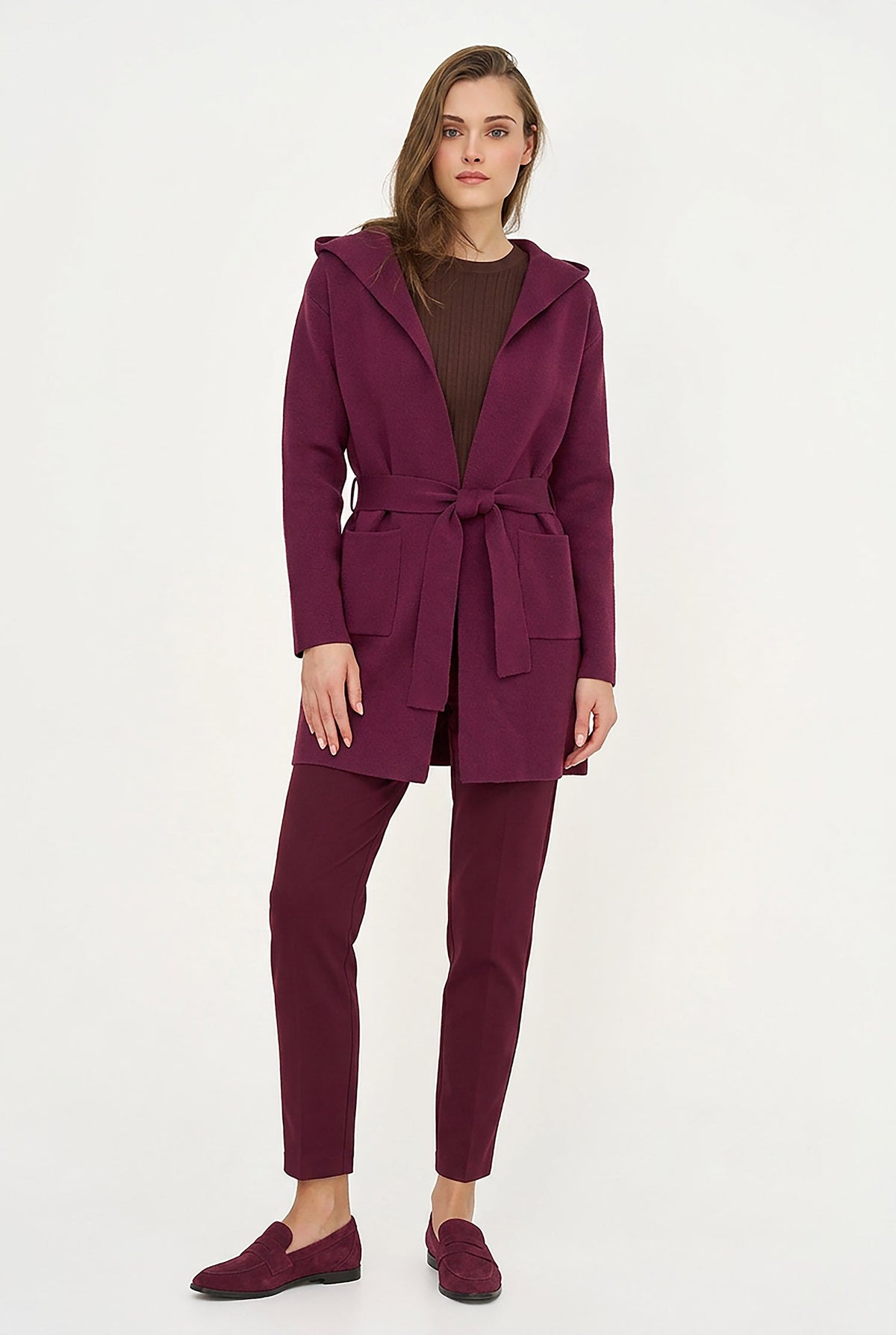 A woman modeling a matching burgundy outfit consisting of a hooded belted coat, slim-fit trousers, and slip-on shoes, against a plain white studio background.