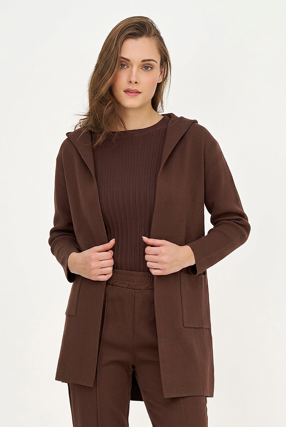 A woman modeling a matching brown knit set consisting of a hooded cardigan, a ribbed long-sleeve top, and trousers, shot against a plain white studio background.