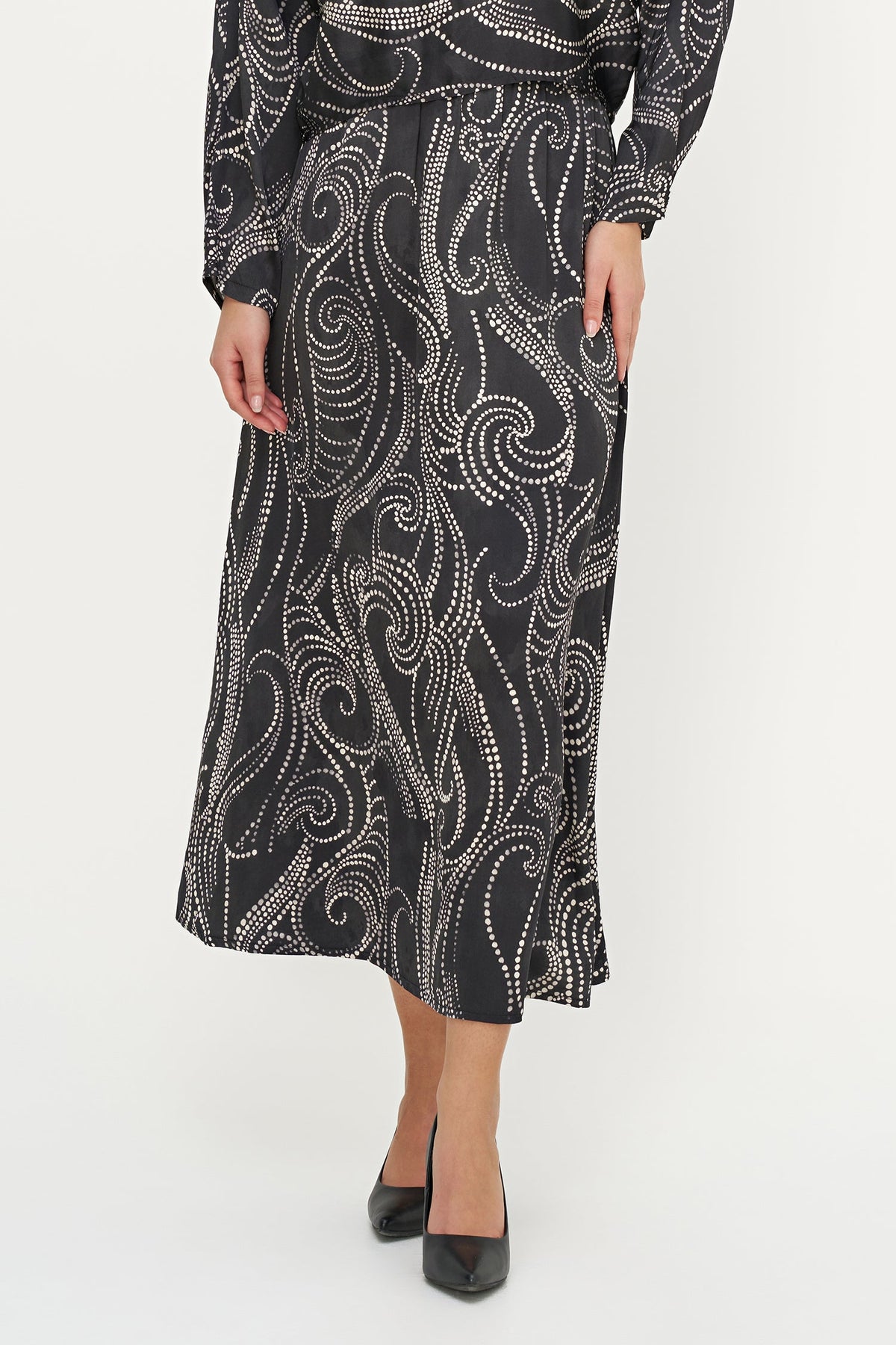 A woman wearing a long-sleeve midi dress with a black base and a white swirling paisley-style pattern, paired with black pointed-toe heels, against a plain white studio background.