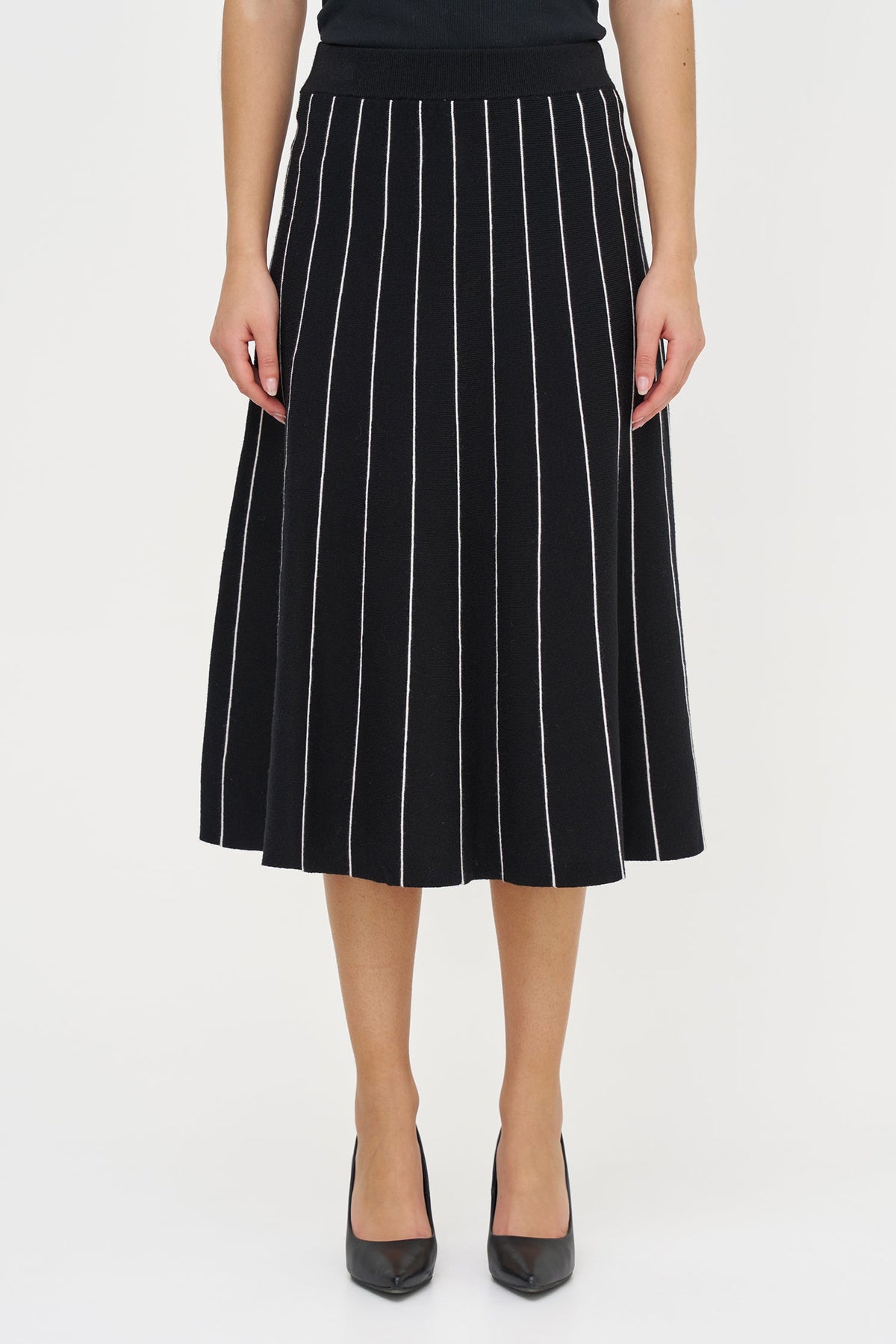 A model wearing a black pleated midi skirt with thin white vertical pinstripes, paired with black pointed-toe heels, against a plain white studio background.