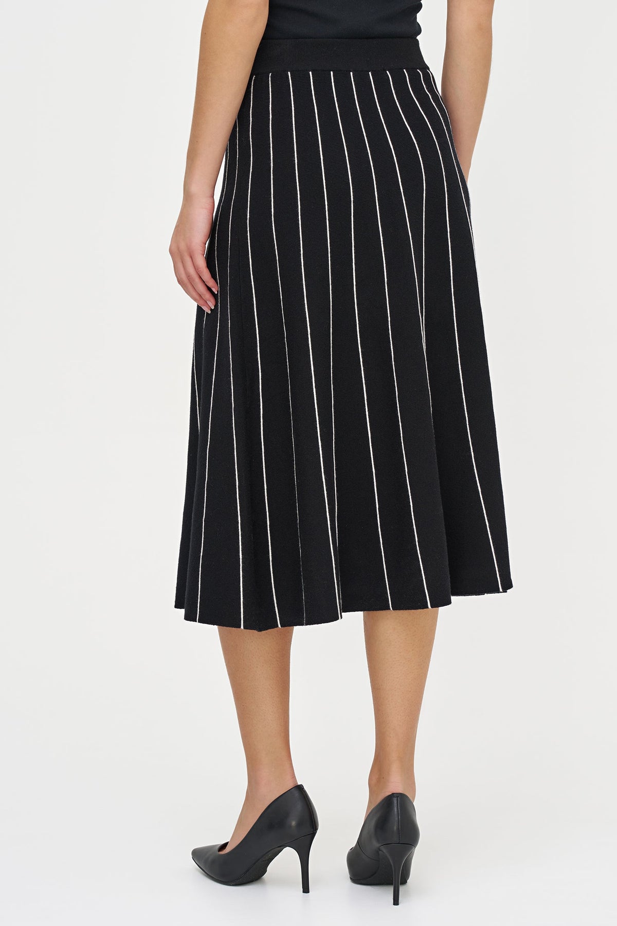 A woman wearing a black midi skirt with thin white vertical pinstripes, paired with black pointed-toe heels, shown from the back against a plain white studio background.