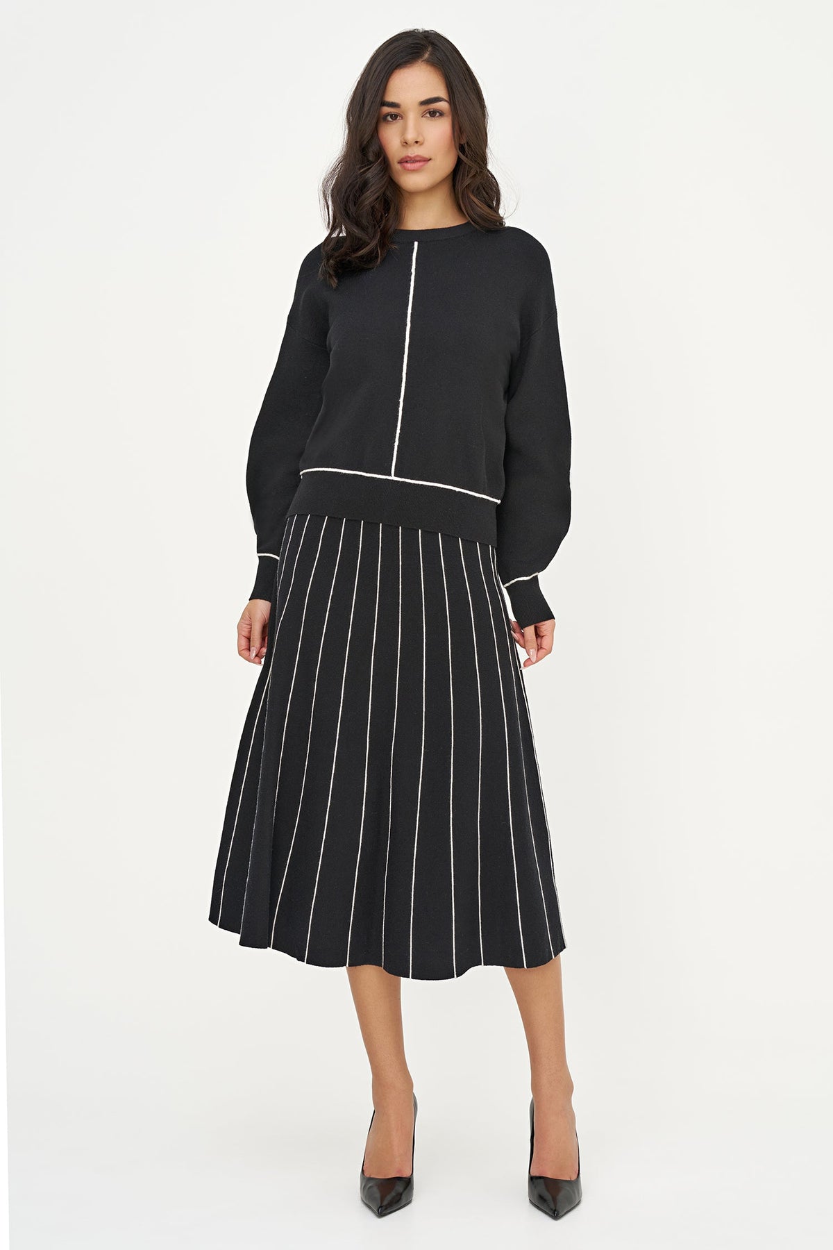 A woman modeling a black long-sleeve top and matching pleated midi skirt set with thin white vertical stripes, paired with black pointed-toe heels, against a plain white studio background.