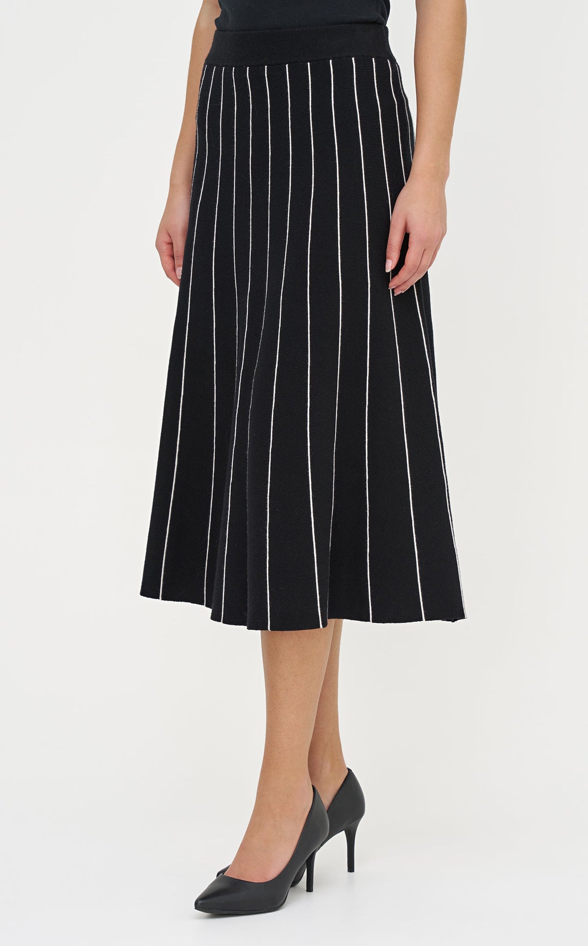 A model wearing a black midi skirt with vertical white stripes, paired with a black top and black pointed-toe heels, shown against a plain white studio background.